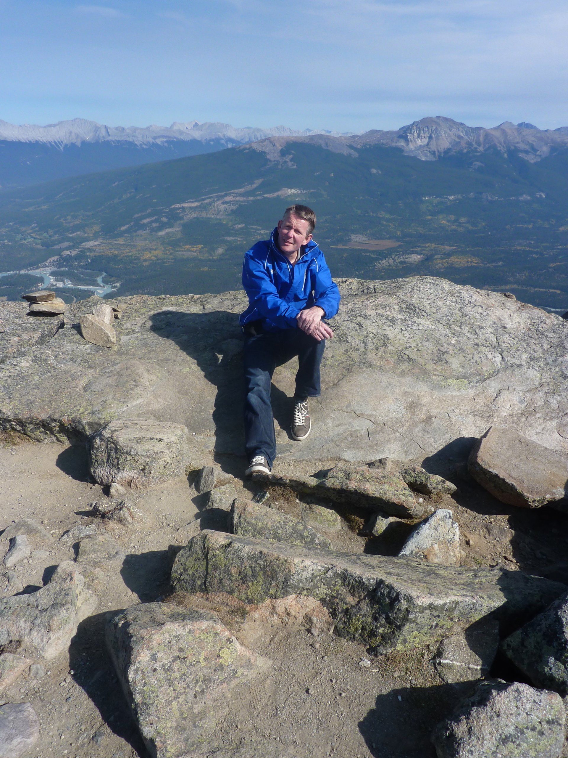 Een man in een blauwe jas zit op een rotsachtige bergtop met uitzicht over een vallei en de bergen in de verte.