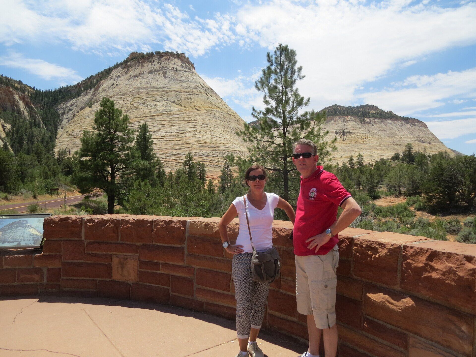 Een stel poseert op een stenen richel met een berglandschap op de achtergrond. Zonnige dag, blauwe lucht.