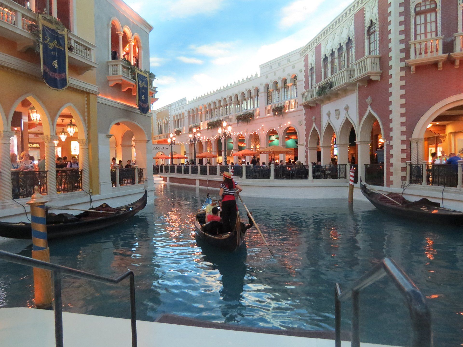 Een gondel op een kanaal in een overdekt gedeelte in Venetiaanse stijl, met kleurrijke gebouwen en mensen die in restaurants dineren.
