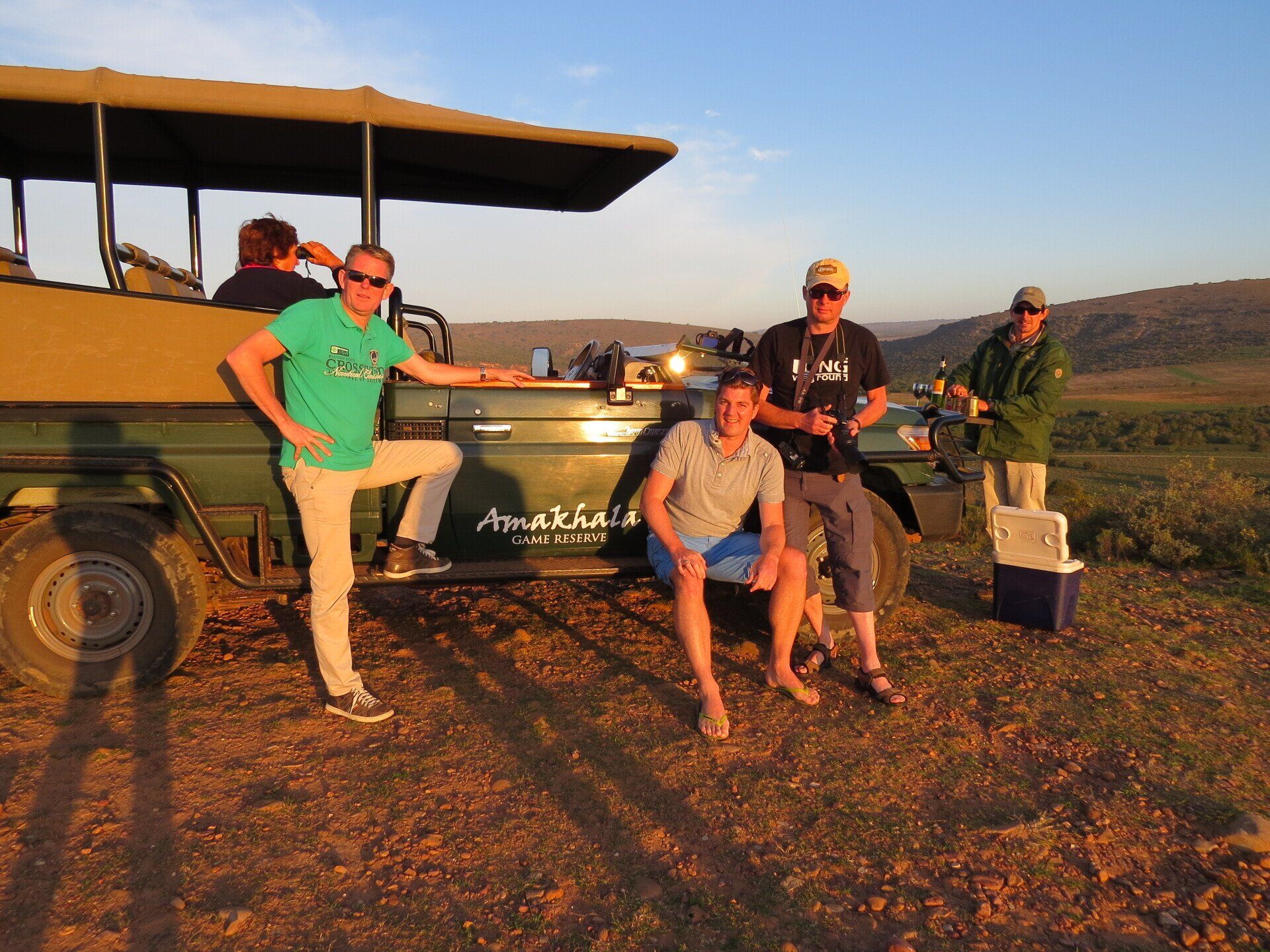 Een groep mensen poseert op een safarivoertuig in een savannelandschap tijdens zonsondergang.