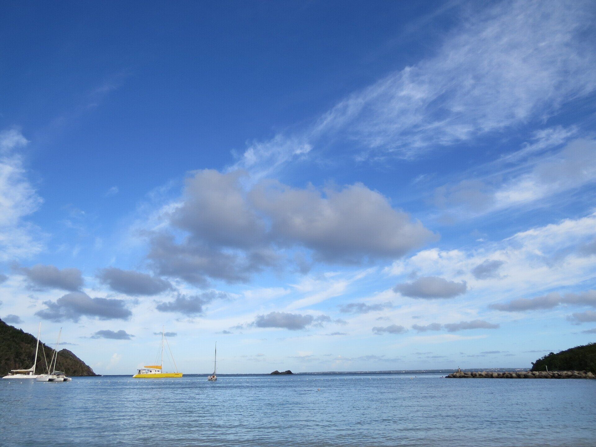Blauwe lucht met ijle, pluizige wolkjes boven een kalme oceaan; een gele zeilboot.