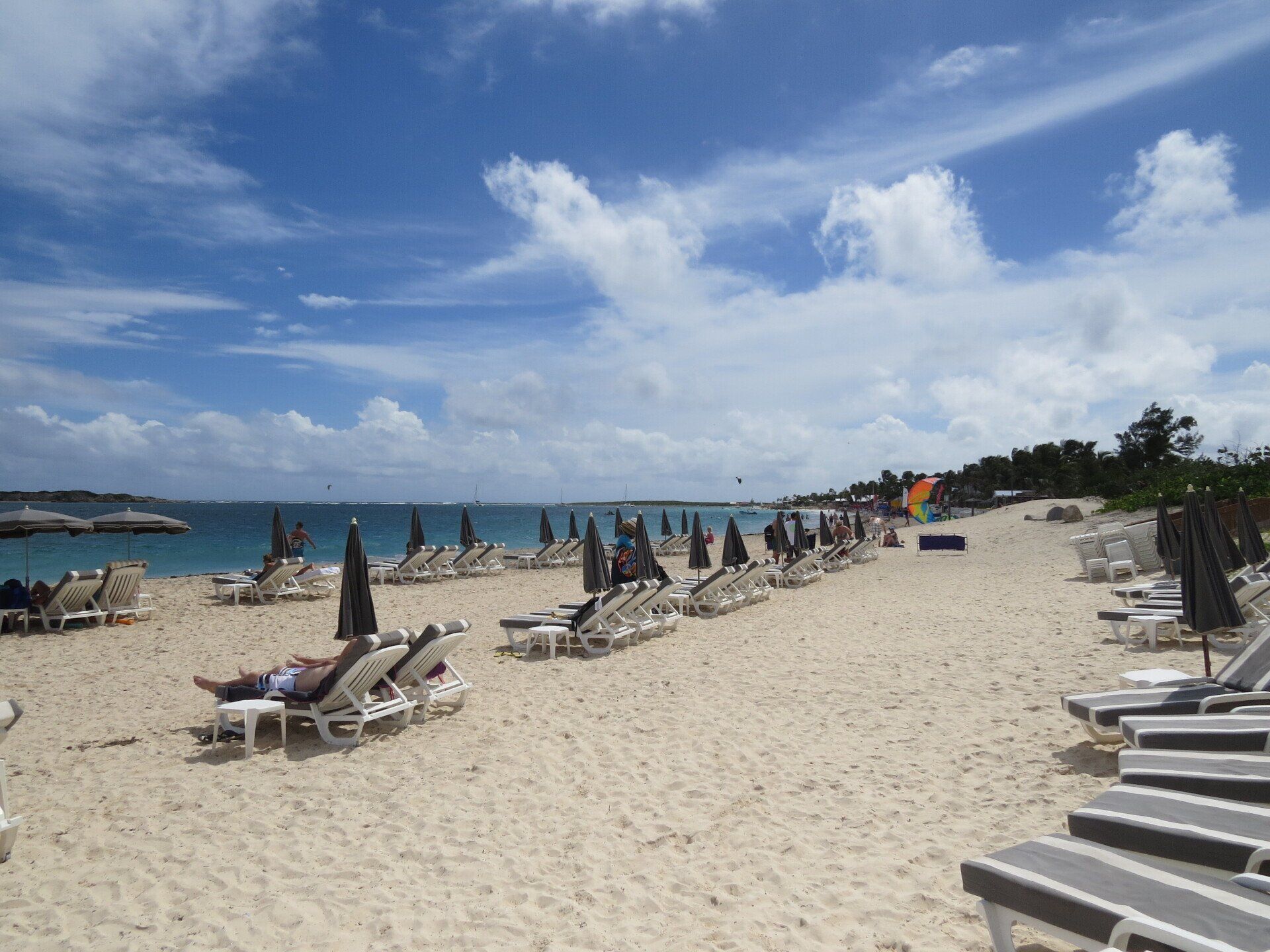Strand met rijen ligstoelen onder parasols, mensen die ontspannen op het zand, blauwe lucht met wolken.