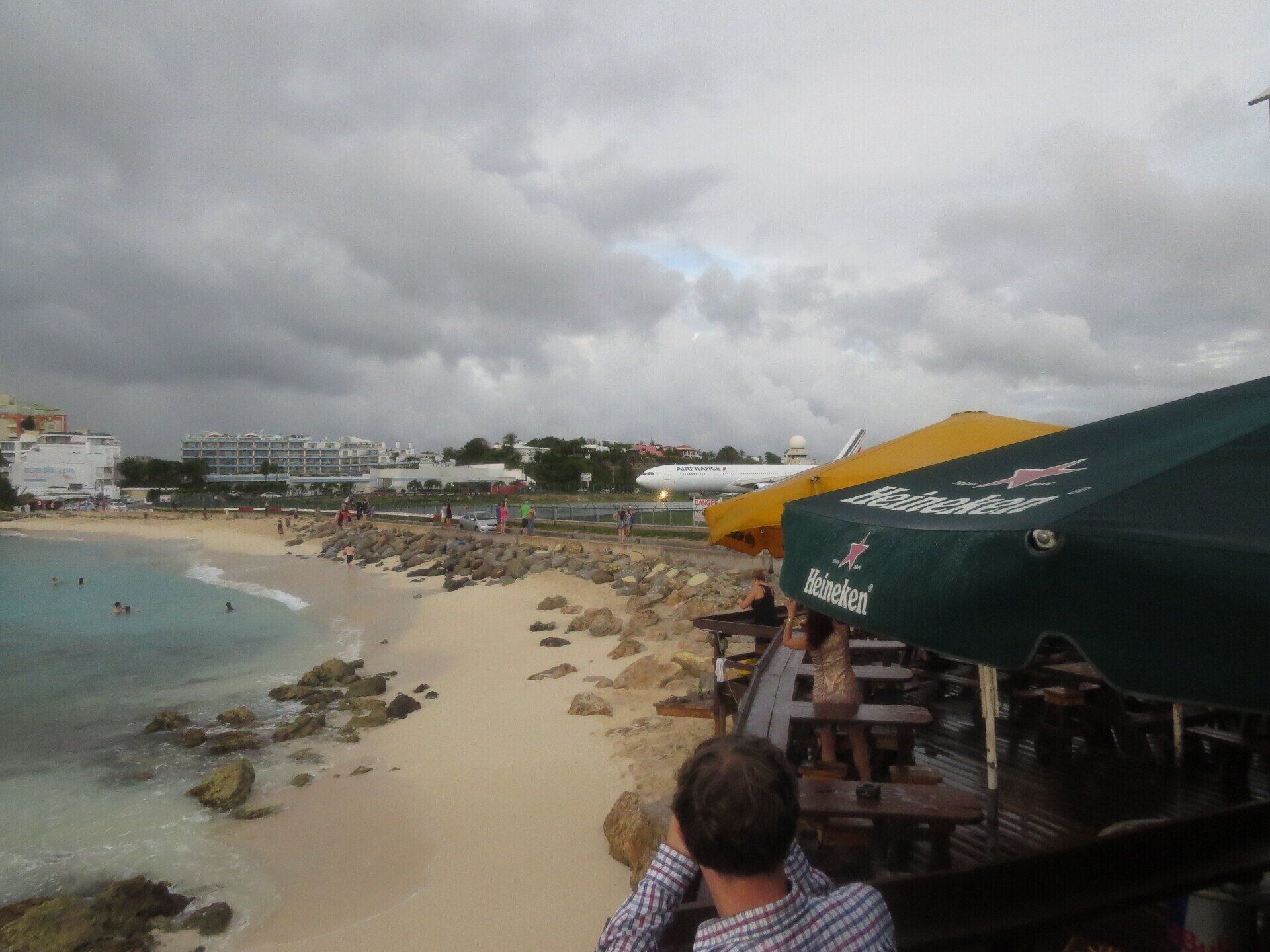 Strandscène met een landend vliegtuig, bewolkte lucht, parasols van een restaurant en mensen op het zand.