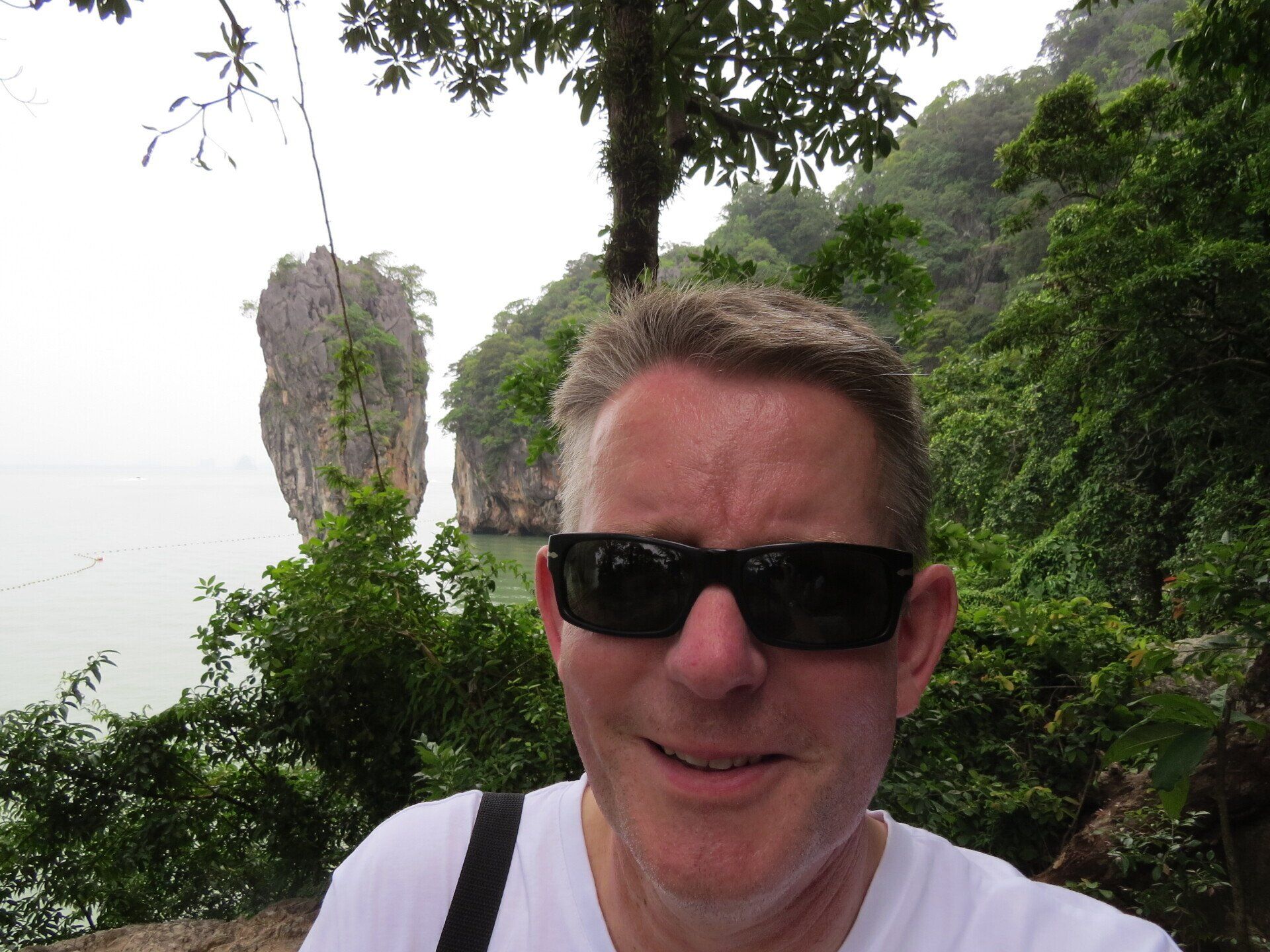 Een man met een zonnebril op, met op de achtergrond een rotsachtig eiland, omgeven door weelderige vegetatie en water.