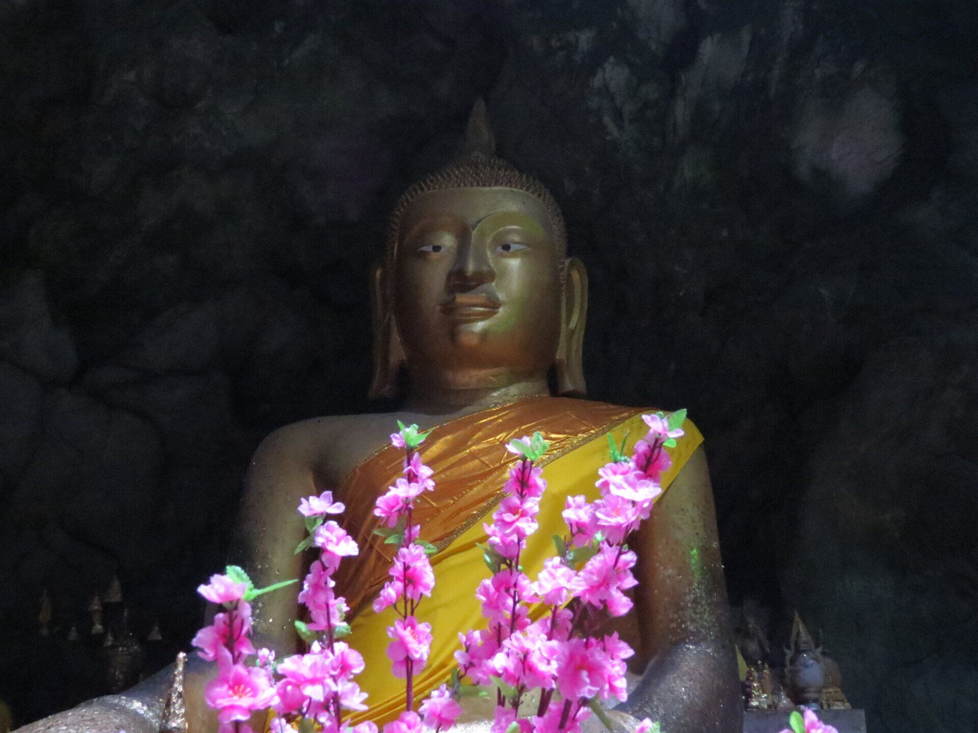 Gouden Boeddhabeeld met roze bloemen, in een donkere grot.