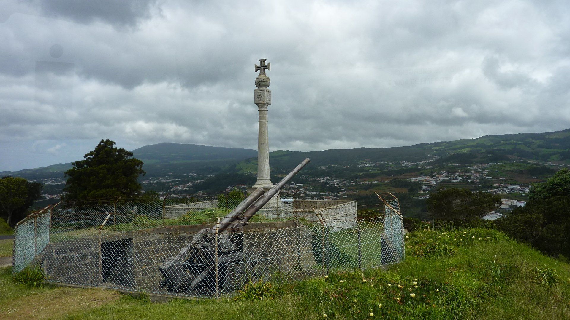 Een stenen kruismonument bovenop een heuvel met uitzicht op een stadje in een vallei, onder een bewolkte hemel.