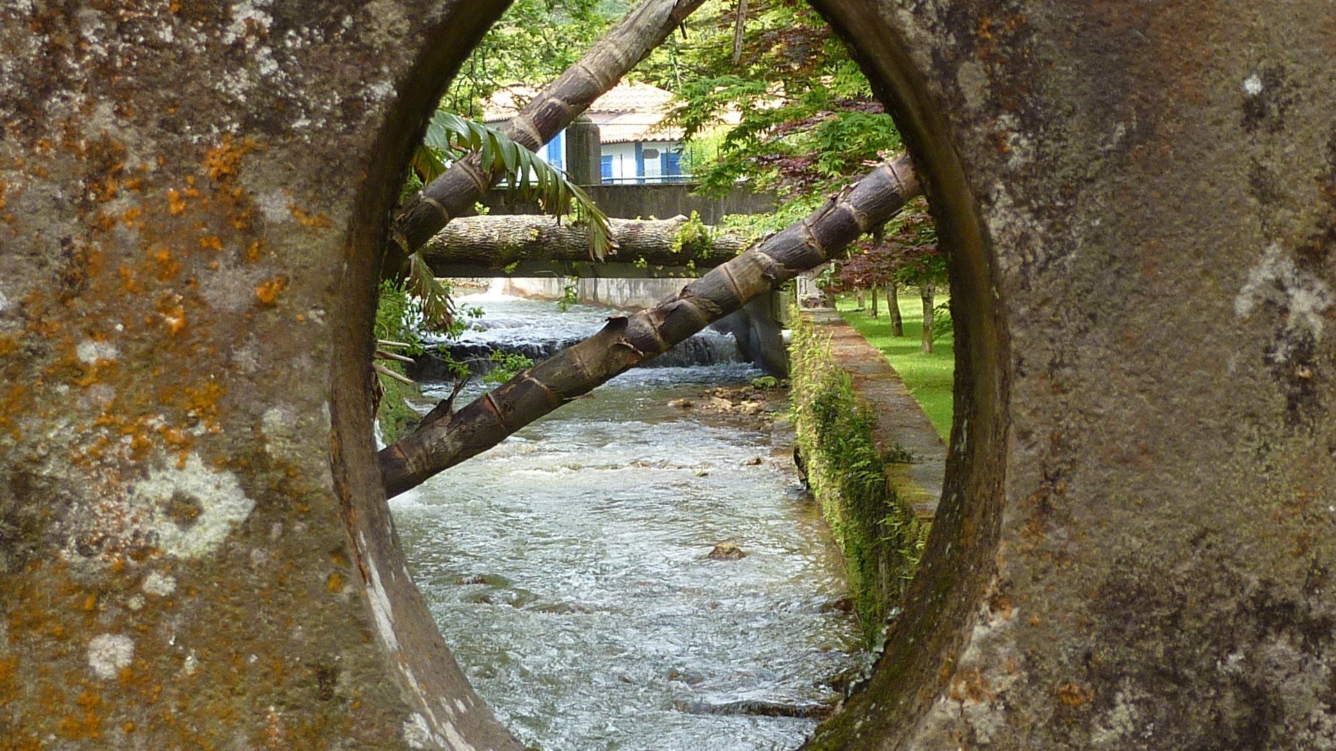 Een stenen muur met een ovale opening die een stromende rivier, bomen en een gebouw in de verte omlijst.