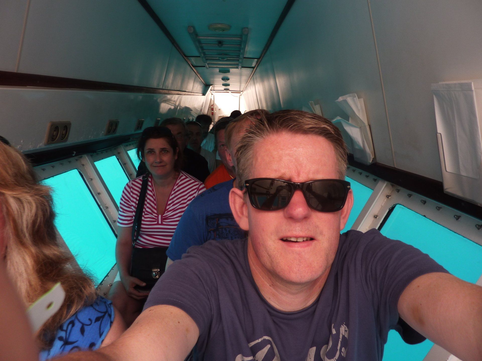 Een man met een zonnebril maakt een selfie in een onderzeeër, terwijl passagiers uitkijken op het turquoise water.