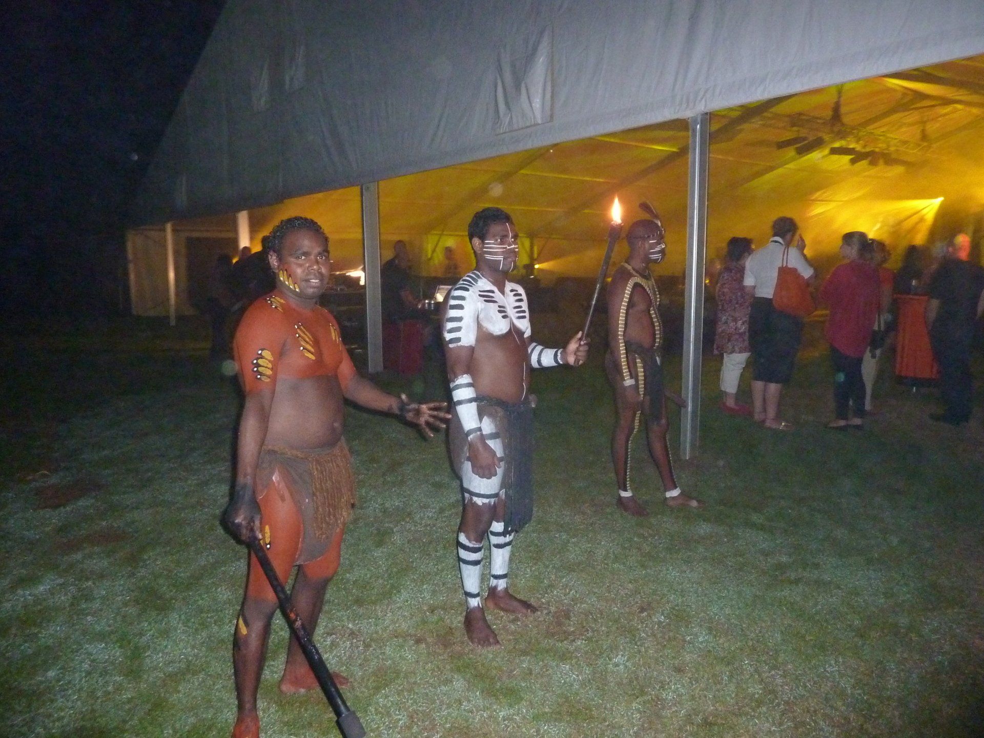 Drie mensen met bodypaint en in traditionele kleding staan ​​met fakkels in hun handen op het gras bij een tent, met een menigte op de achtergrond.