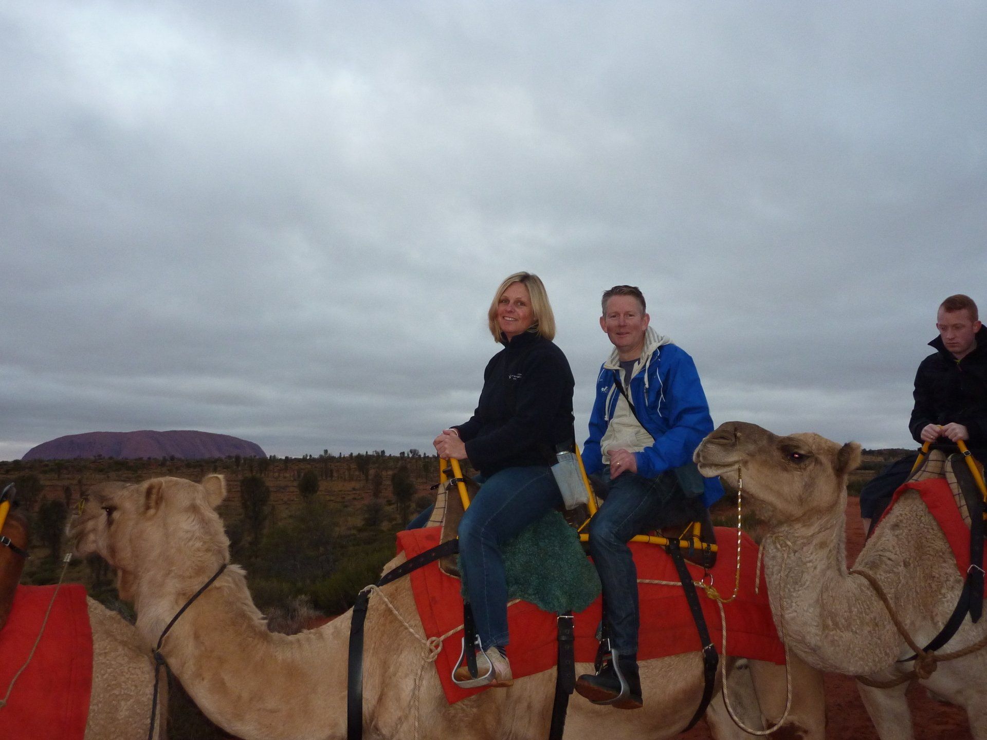 Mensen rijden op kamelen in een woestijnlandschap, met Uluru in de verte zichtbaar onder een bewolkte hemel.