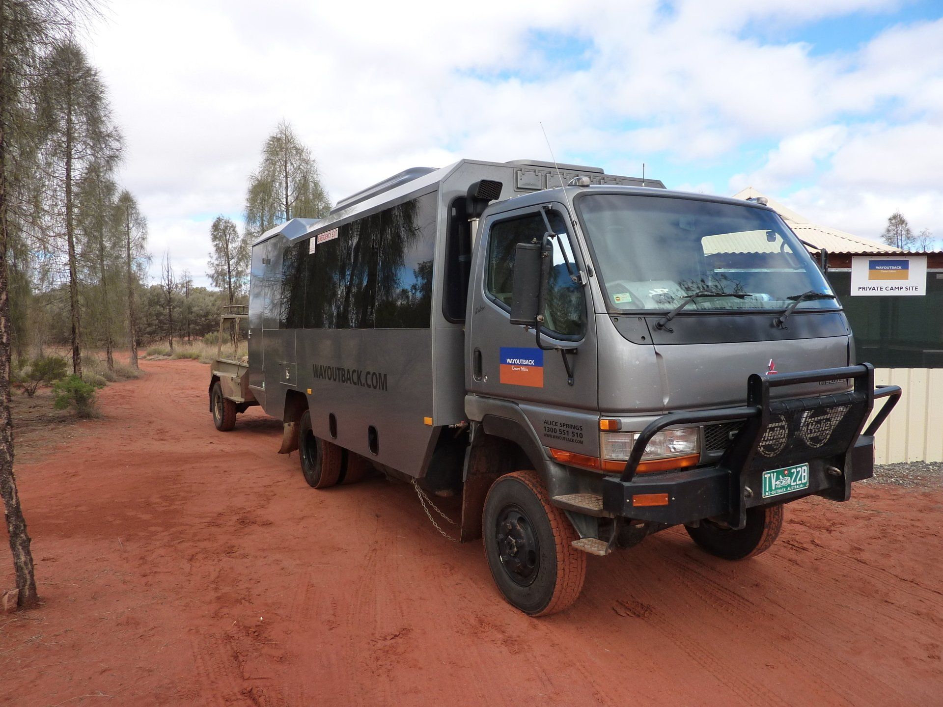 Grijze terreinwagen met camper op een rode zandweg. Op de achtergrond zijn bomen en een klein gebouw te zien.