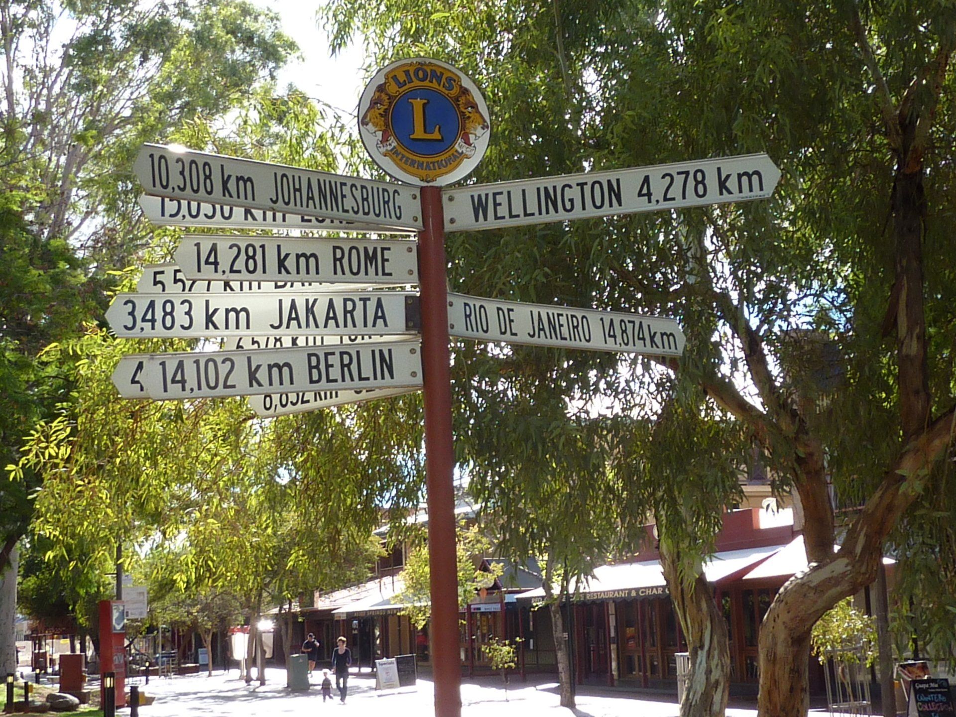 Wegwijzer met afstanden naar verschillende steden, waaronder Wellington, Jakarta en Berlijn, met een Lions Club-embleem.