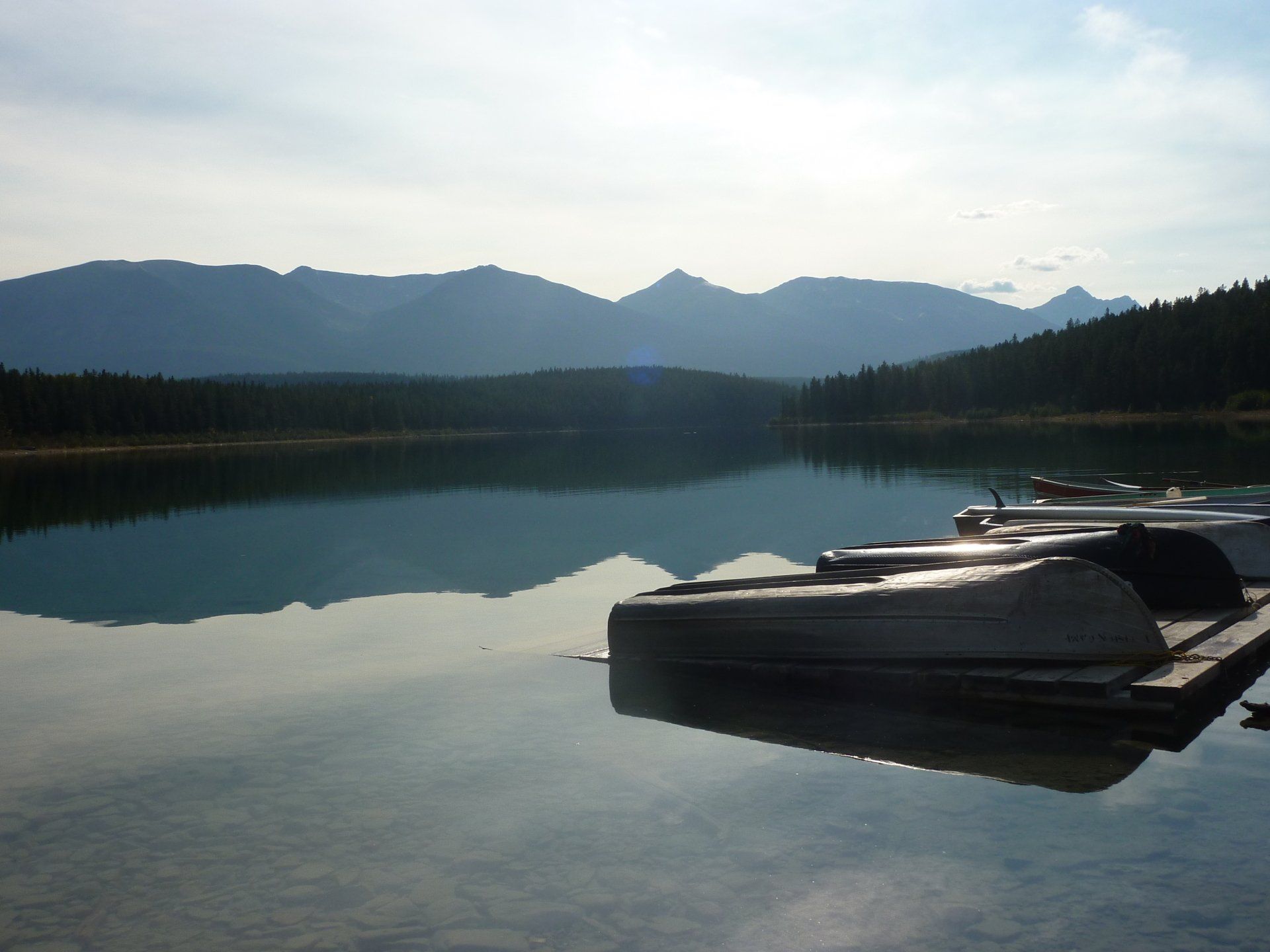 Een tafereel aan een meer met boten aan de steiger en bergen in de verte die weerspiegelen in het stille water.