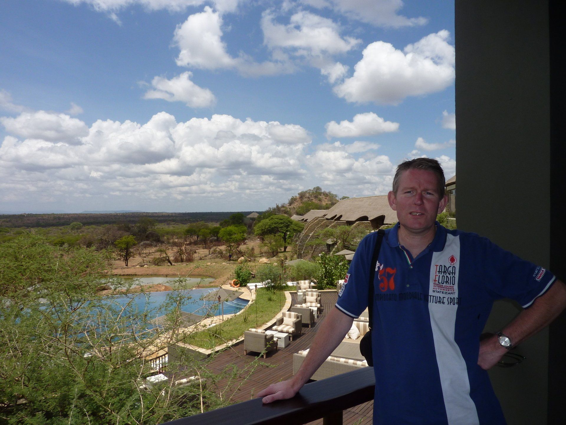 Man op balkon met uitzicht op een zwembad en landschap onder een bewolkte hemel.