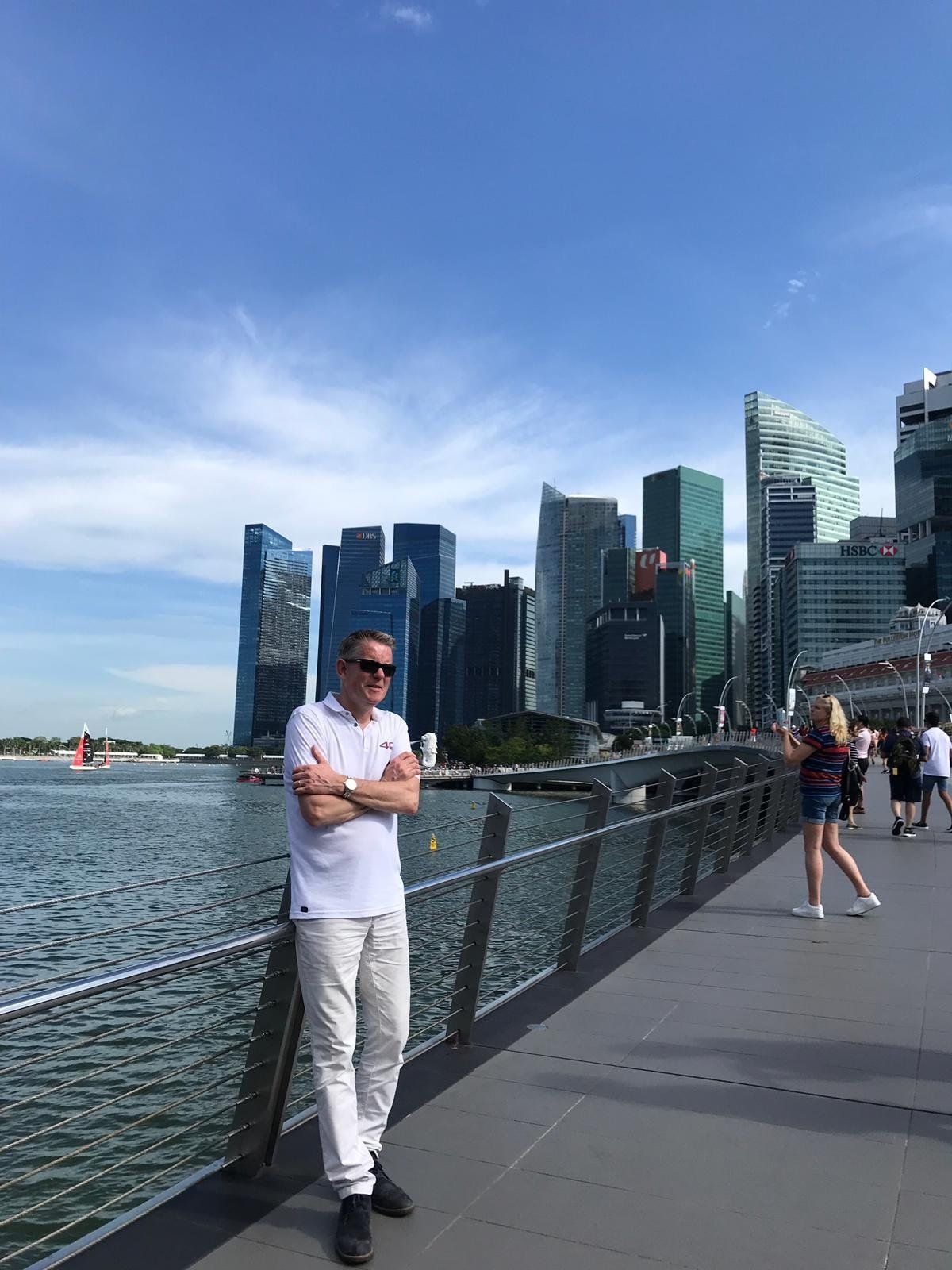 Man met gekruiste armen op een wandelpromenade langs het water met de skyline van de stad op de achtergrond.