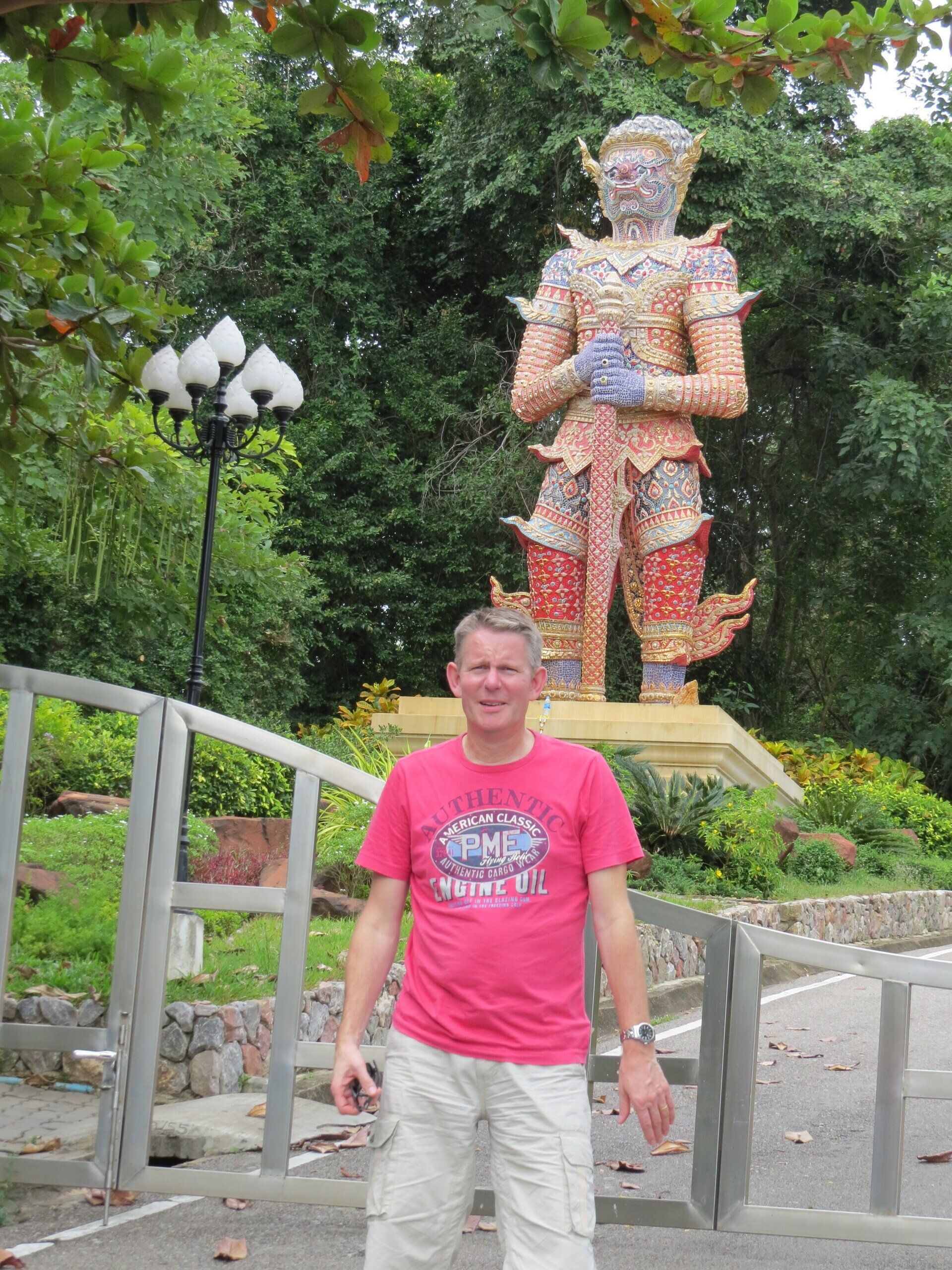 Een man in een roze shirt staat voor een groot, sierlijk standbeeld; op de achtergrond zijn bomen en een lantaarnpaal te zien.