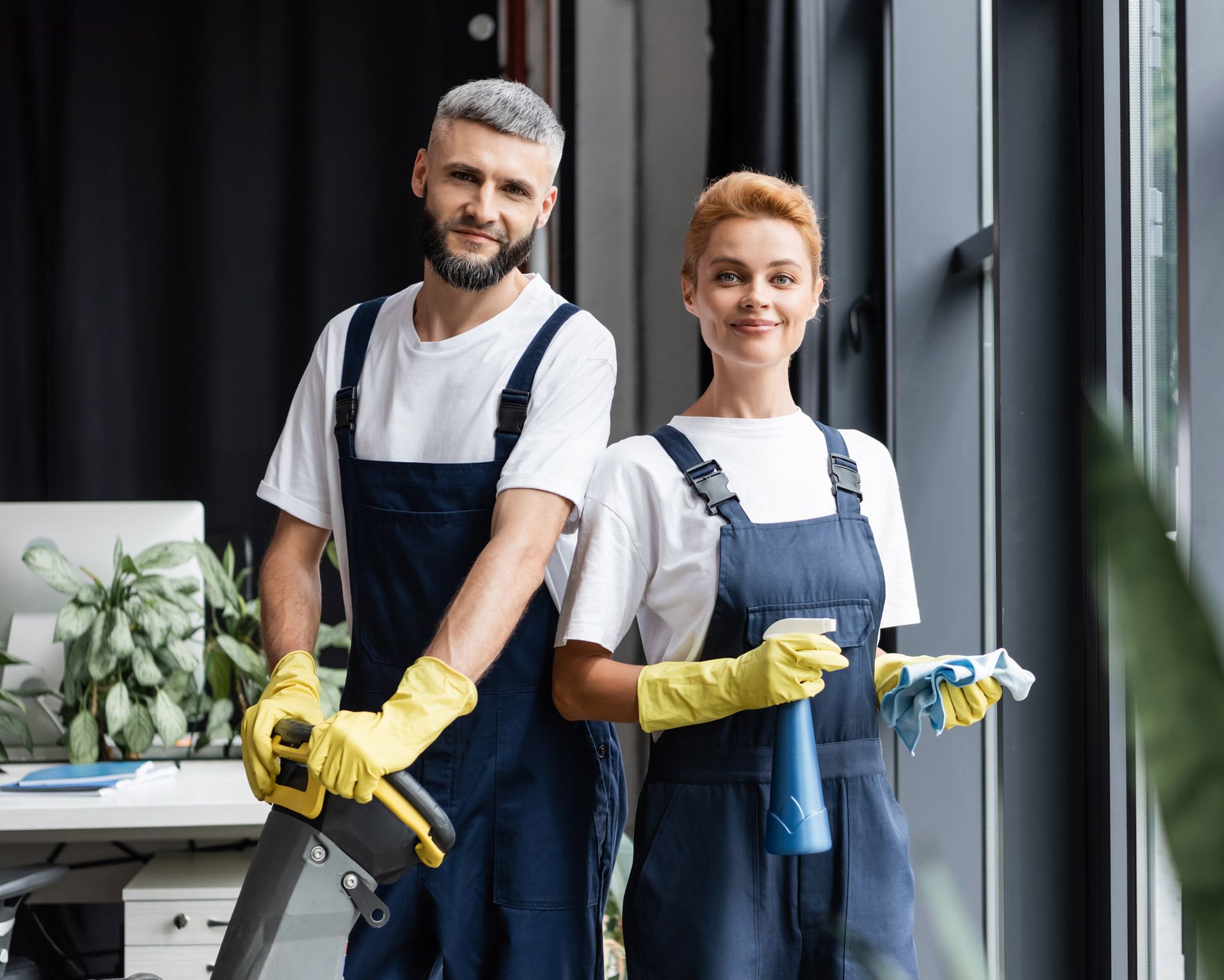 A man and a woman are standing next to each other in an office holding cleaning supplies.