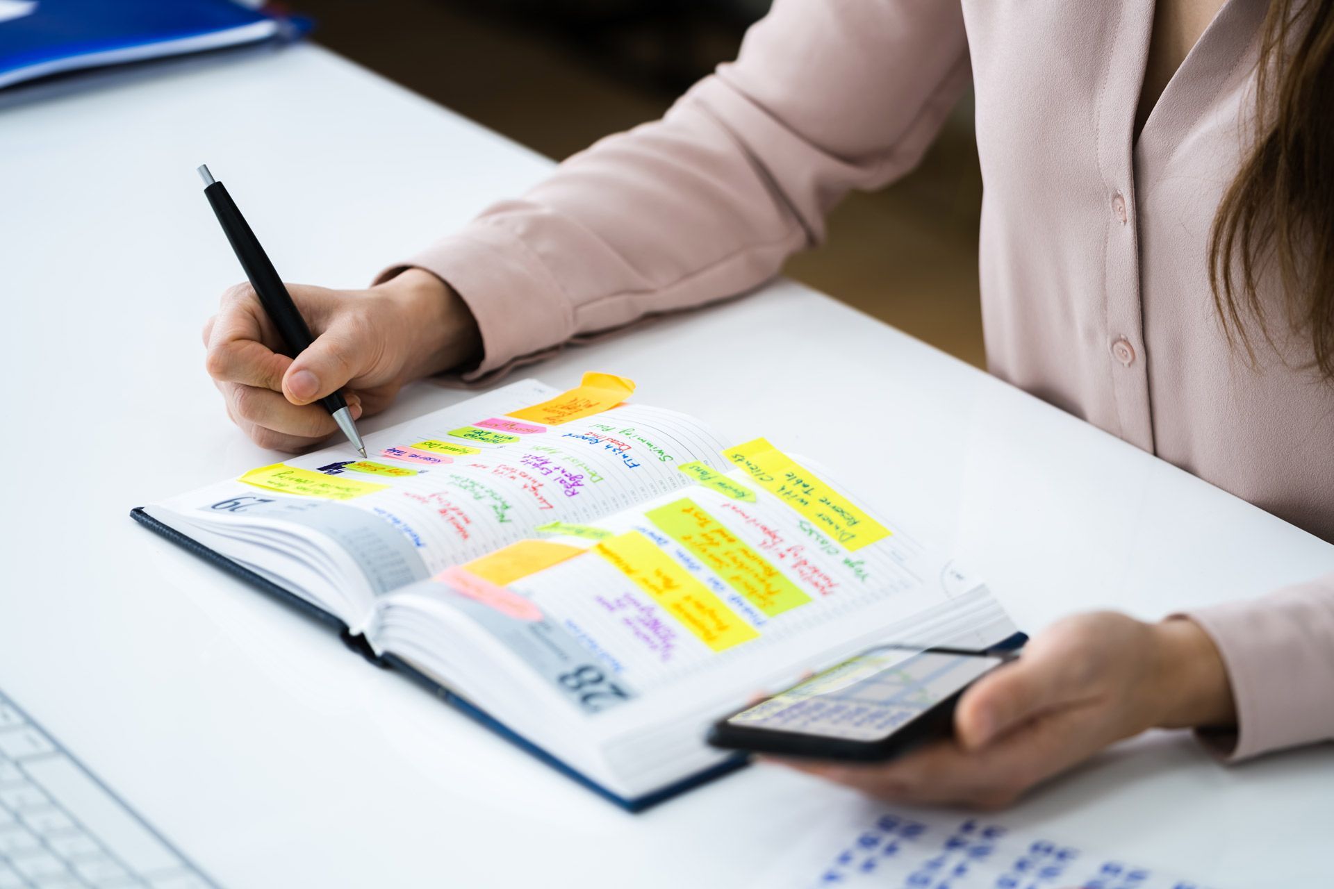 A woman is sitting at a desk writing in a notebook while holding a cell phone