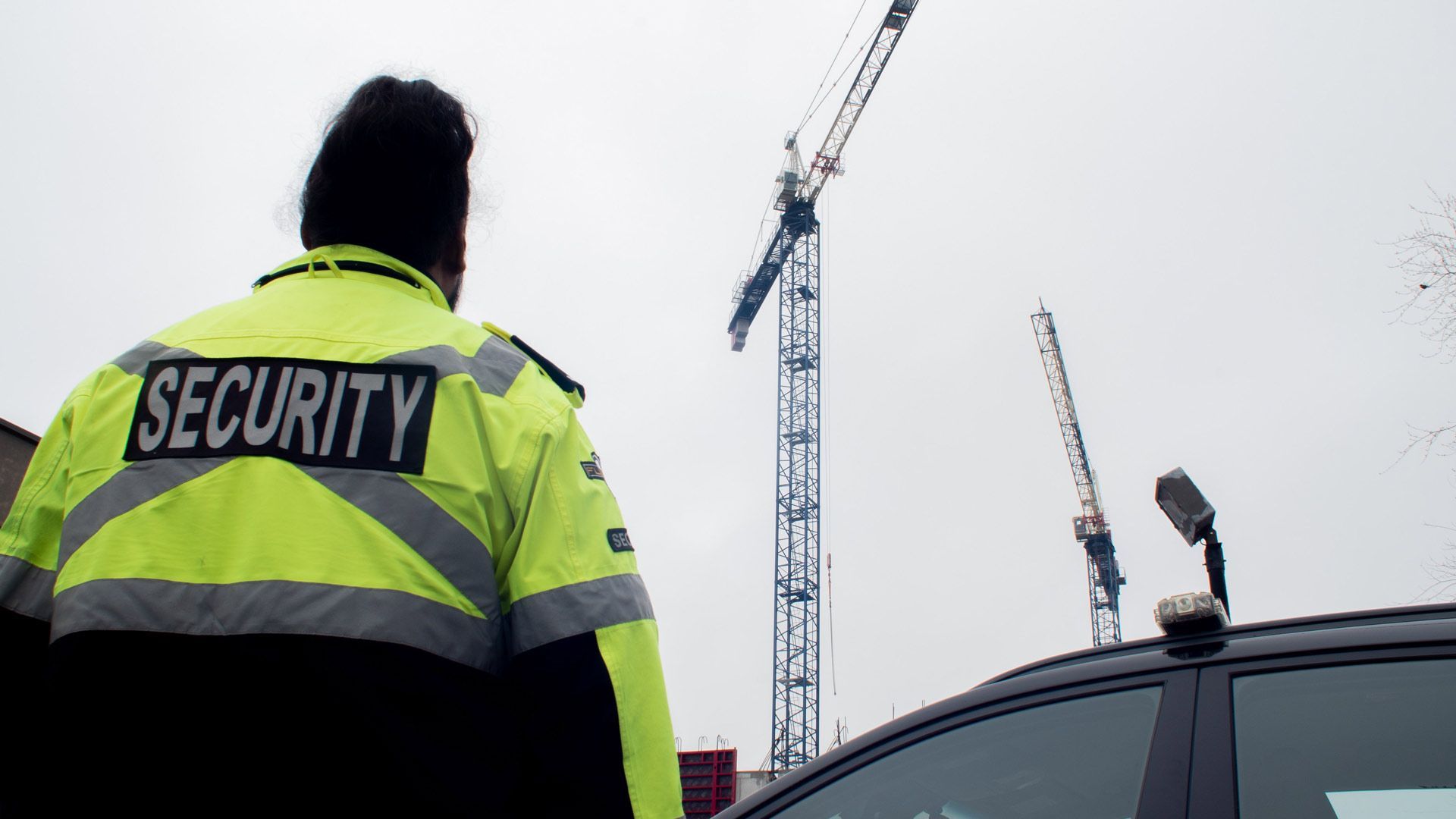 A security guard is standing next to a car and looking at a crane.