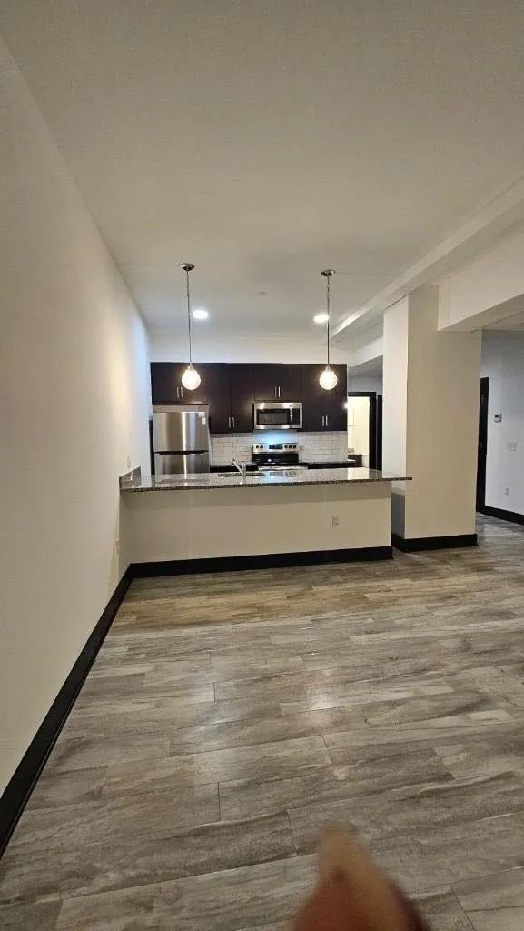 A person is standing in an empty room with a kitchen in the background.