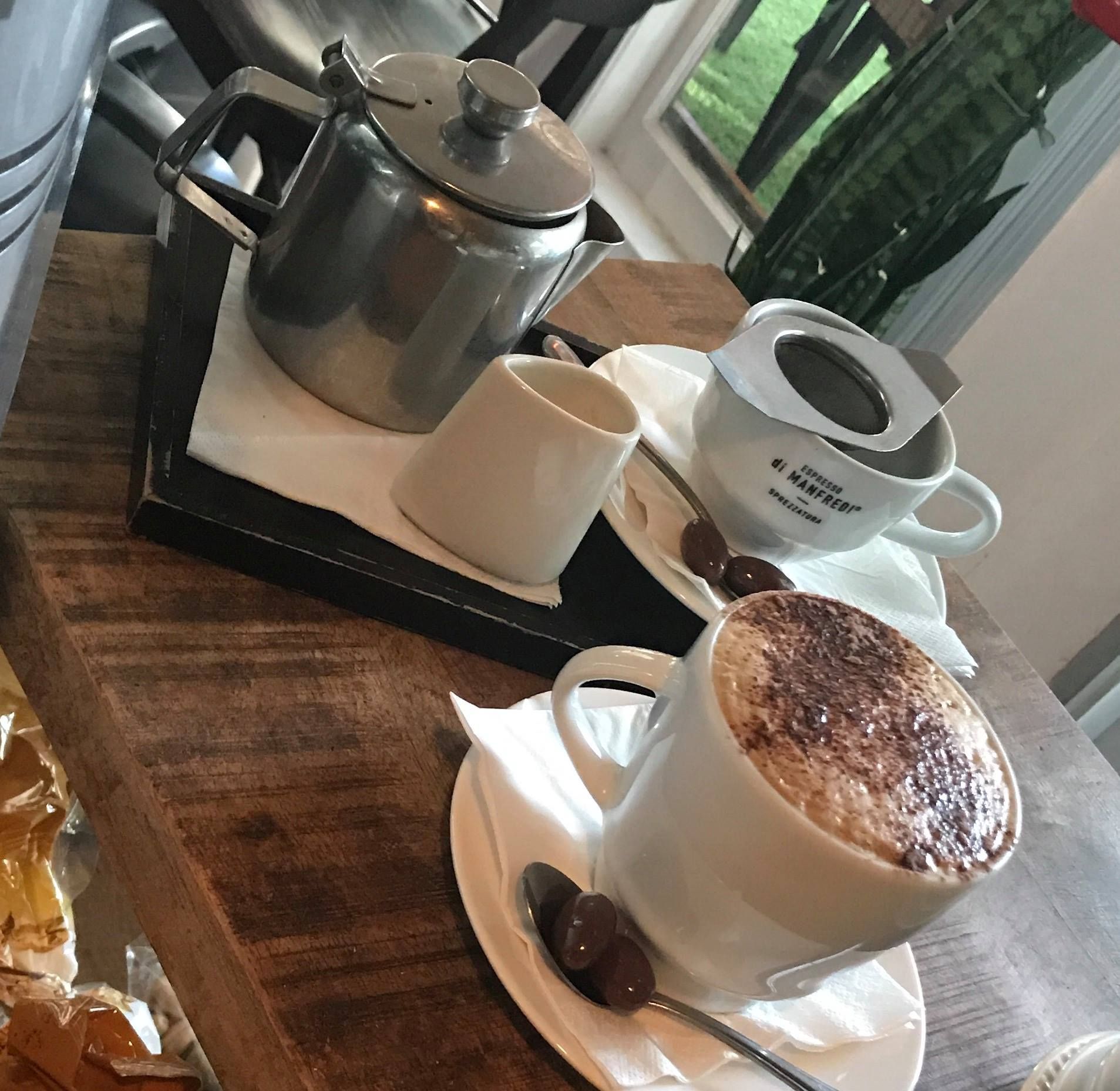 Cappuccino and tea set on a wooden table; white cups and saucers, silver teapot, chocolate decorations.