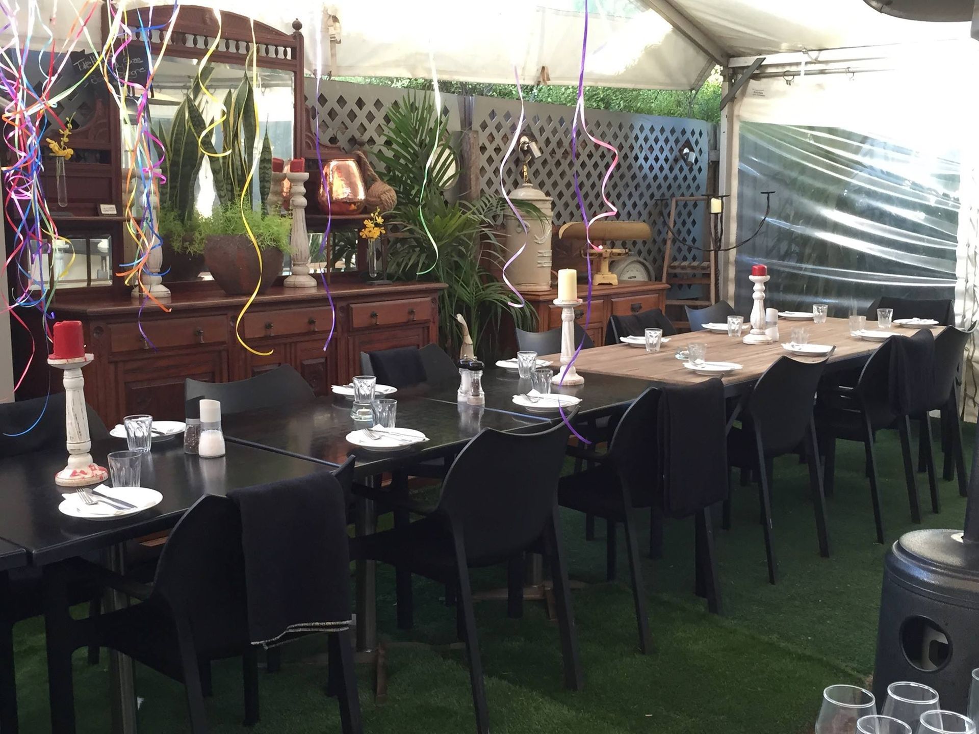 Outdoor dining area with tables set for a meal, under a tent. Black chairs and decorative sideboards are present.