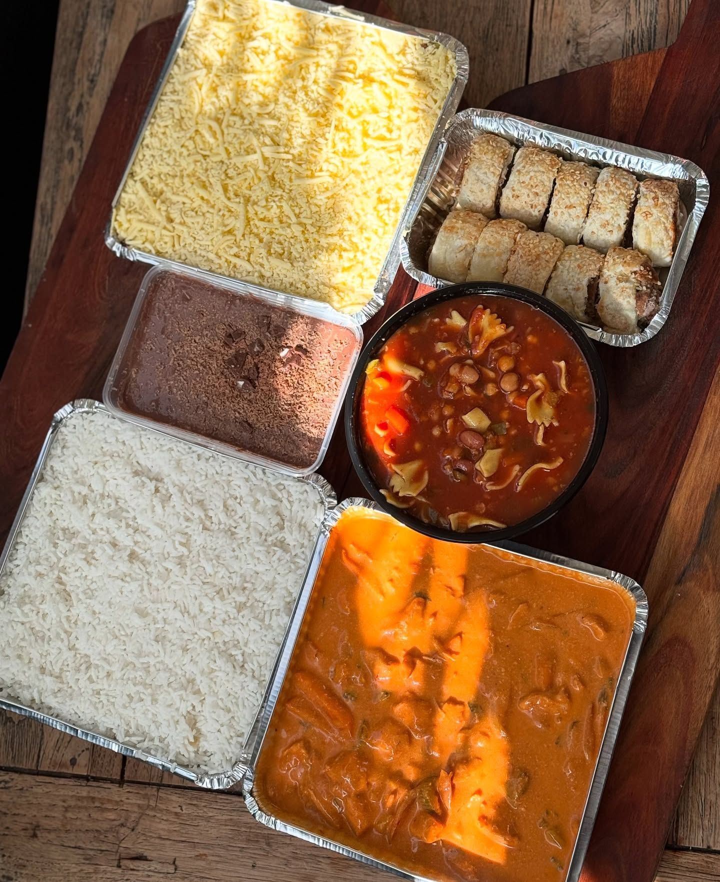 Containers of prepared food: rice, cheese, beans, sauce, bread rolls, and meat in a rich sauce, on a wooden surface.