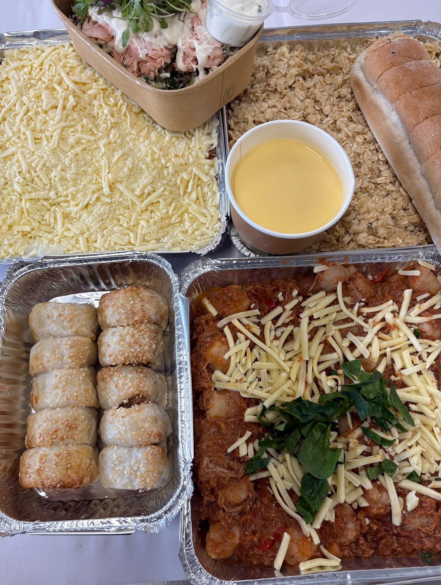 An assortment of prepared food in foil containers, including rolls, casserole, and salad, cheese and bread.