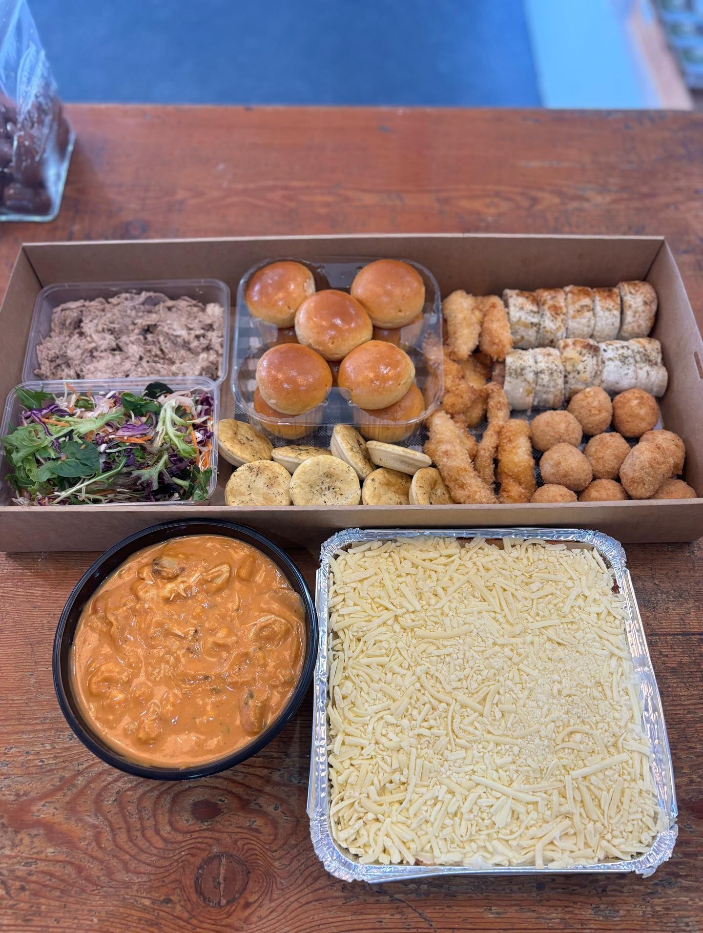 Boxed catering spread with various food items: rolls, sushi, salad, curry, and casserole.