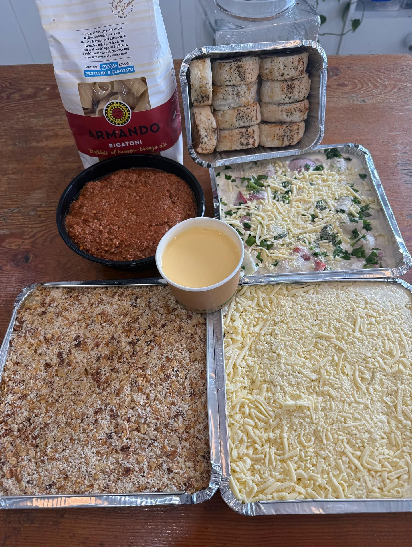 Prepared meal components in foil trays, including bread, sauce, and grated cheese, on a wooden surface.