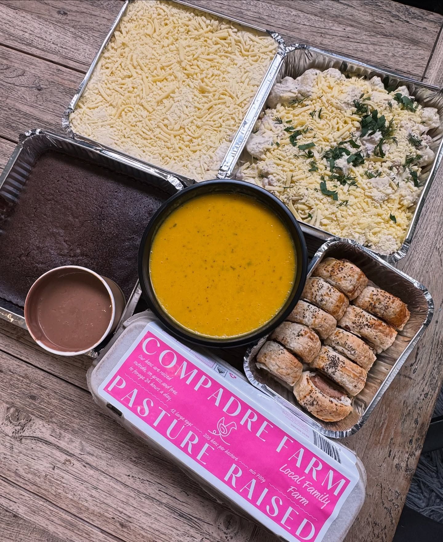 A spread of prepared meals: cheesy casserole, cake, soup, rolls, eggs, and chocolate sauce.