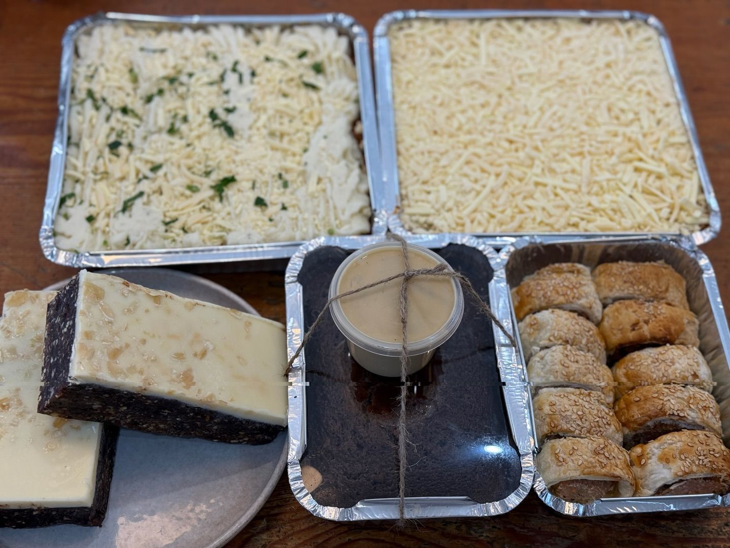 Aluminum trays of prepared food: lasagna, cheese, brownies, dip, and sausage rolls on a wooden surface.