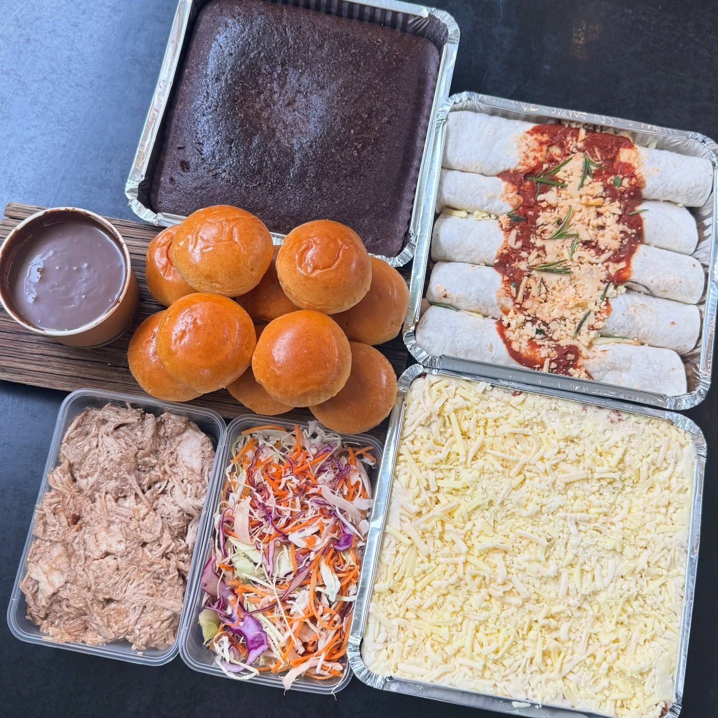 An assortment of food in foil containers, including cake, rolls, shredded meat, slaw, casserole, and enchiladas.