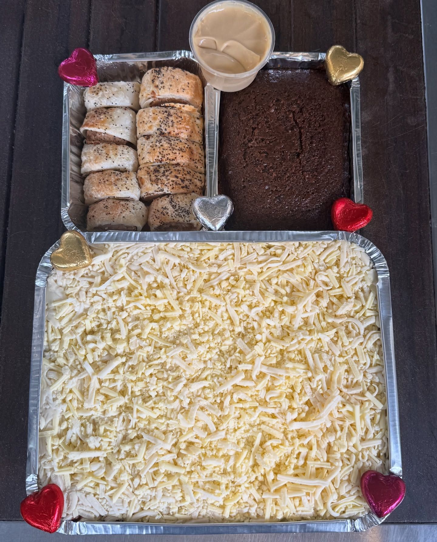 Two foil containers with food. One has shredded cheese, the other has rolls and cake with dip.
