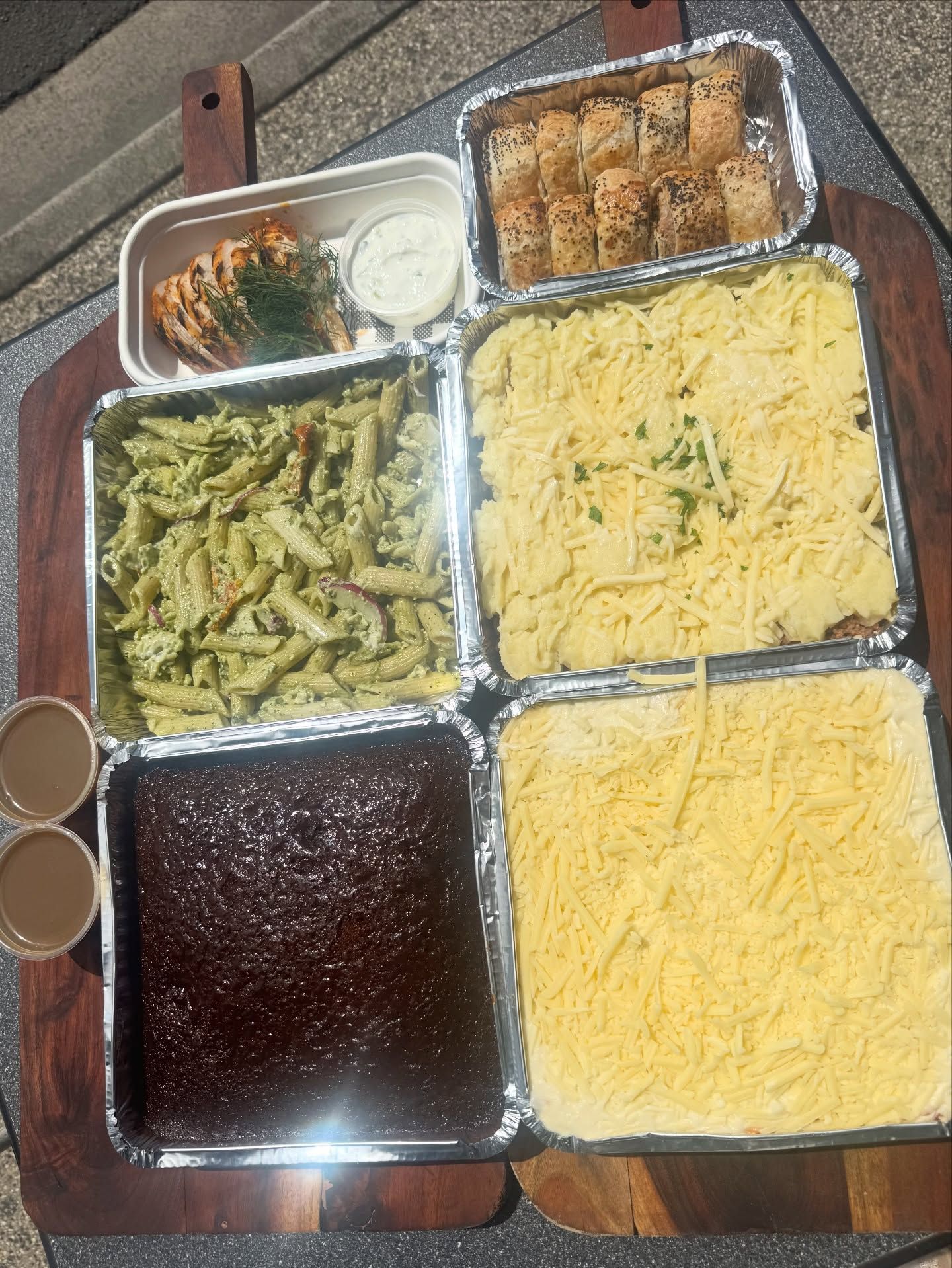 Takeout meal with pasta, chicken, baked dishes, garlic bread, and brownies in aluminum containers on a wooden board.