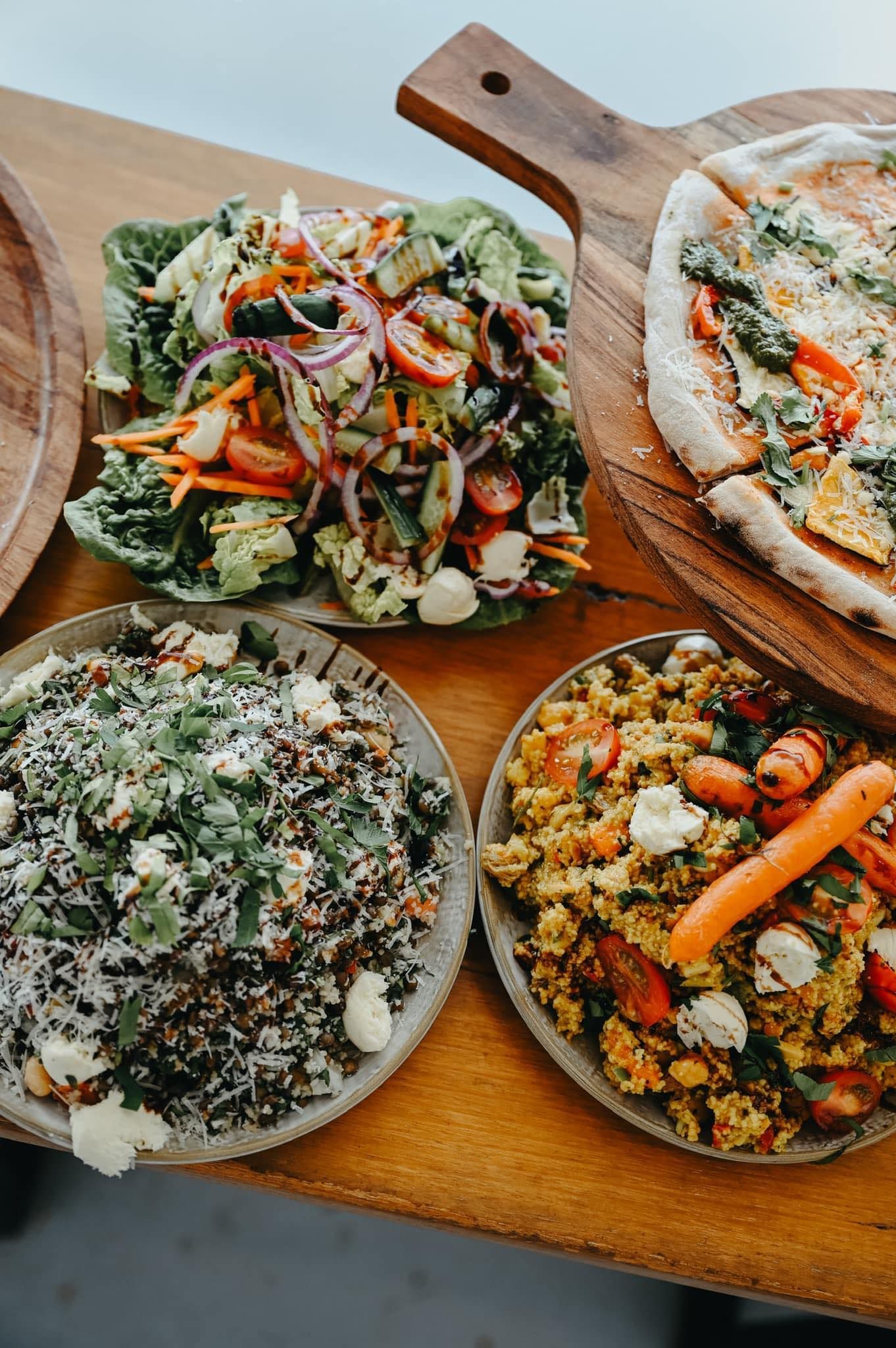 Overhead shot of a wooden table with salads, pizza and various veggie dishes.