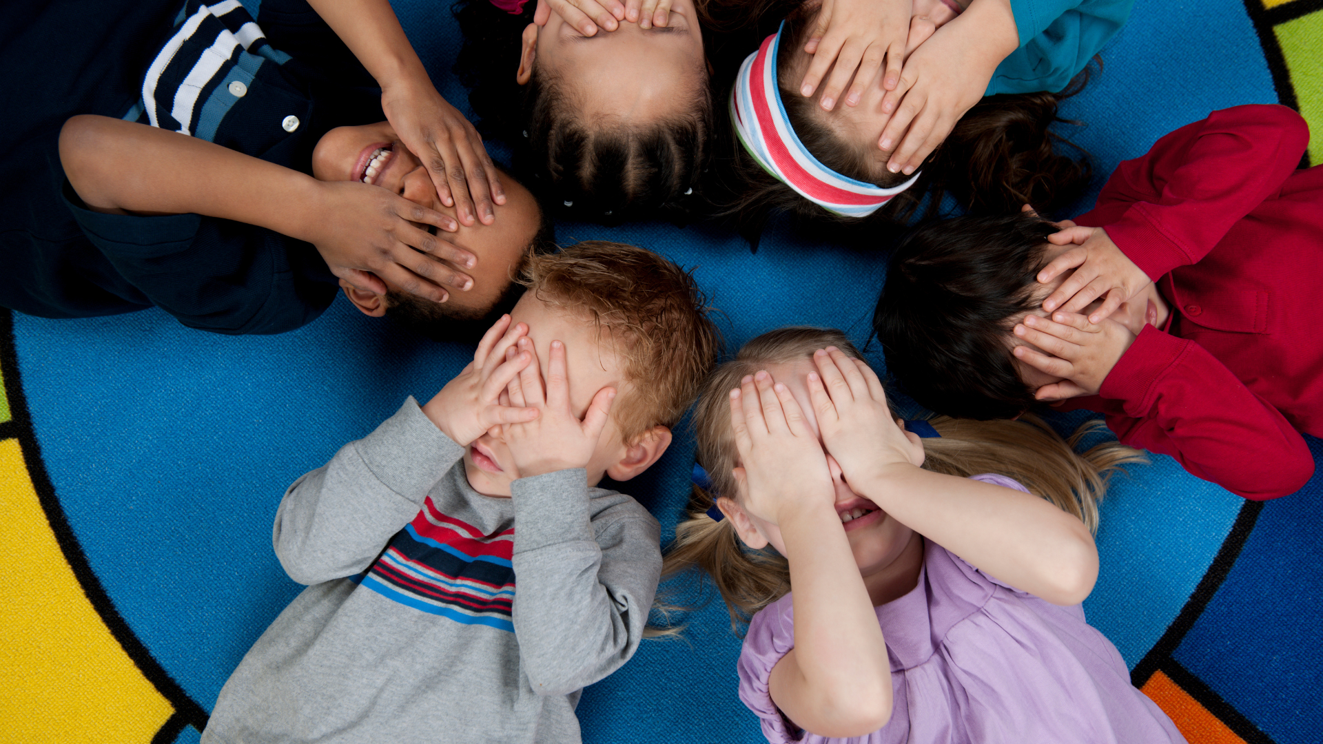 A group of children are laying in a circle covering their eyes with their hands.