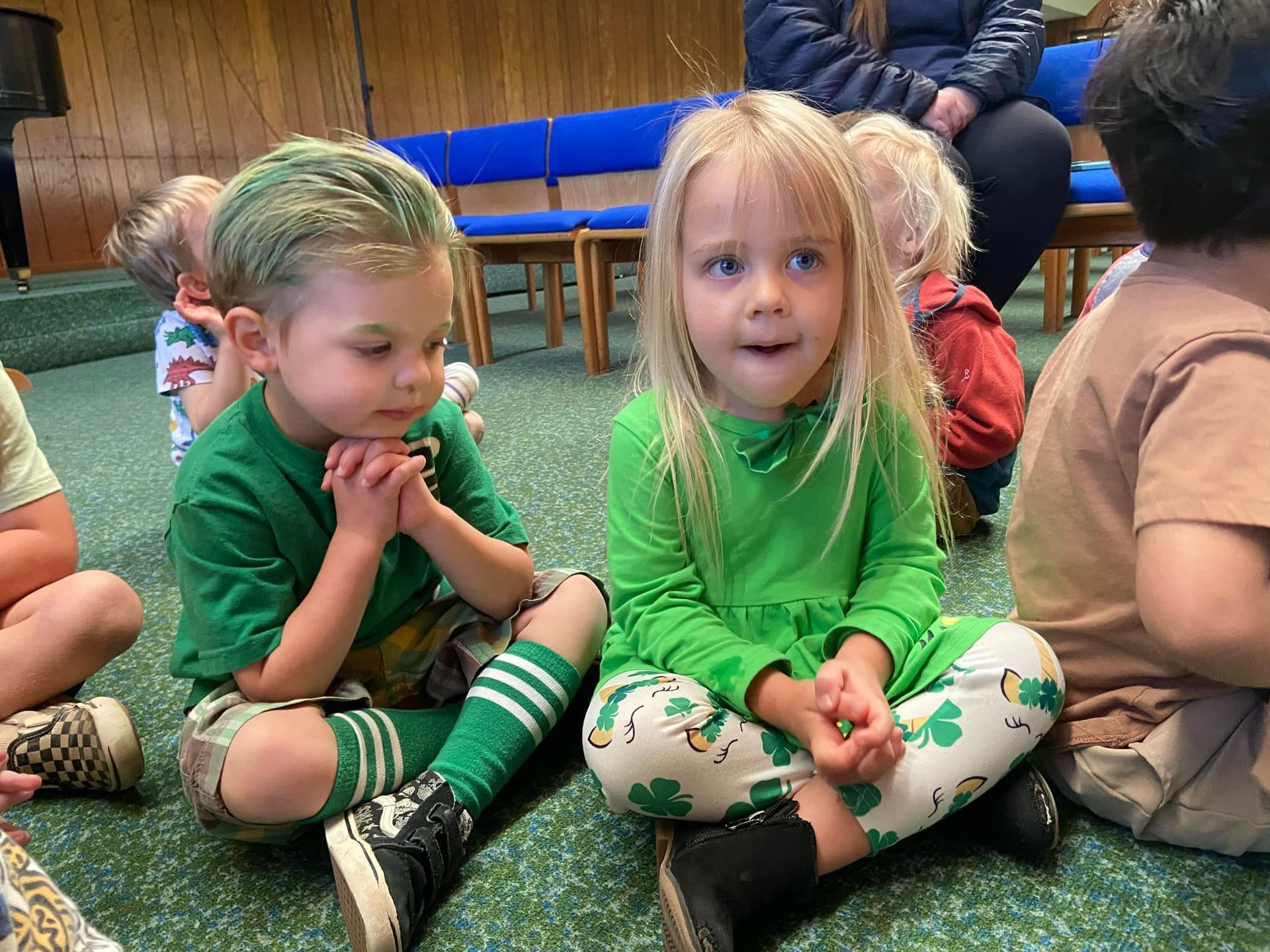 A group of children are sitting on the floor wearing green clothes.