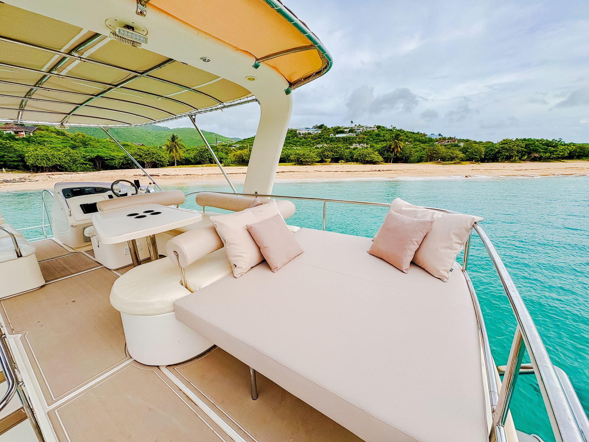 Boat deck with beige sunbed, pillows, and a white table, overlooking turquoise water and beach.