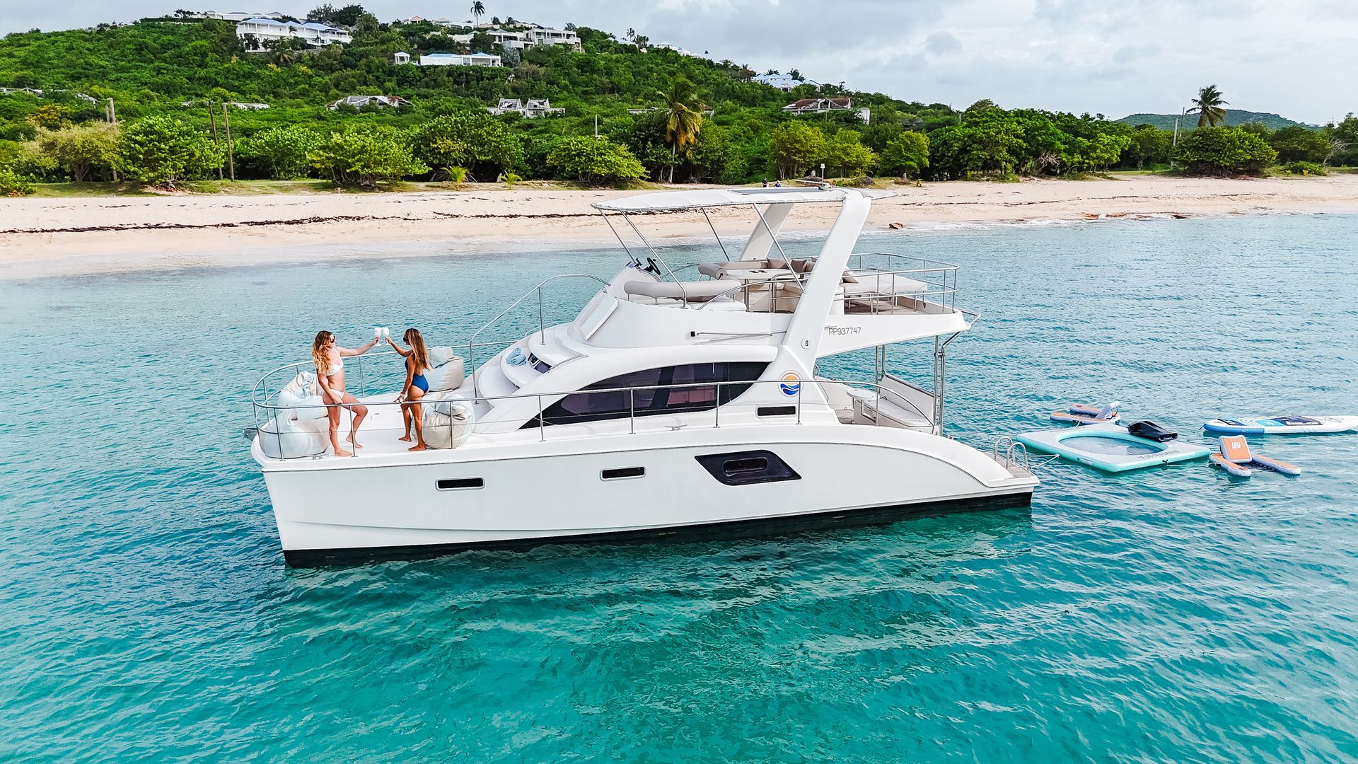 White catamaran in turquoise water with two people standing on the deck. Beach and green hills in background.
