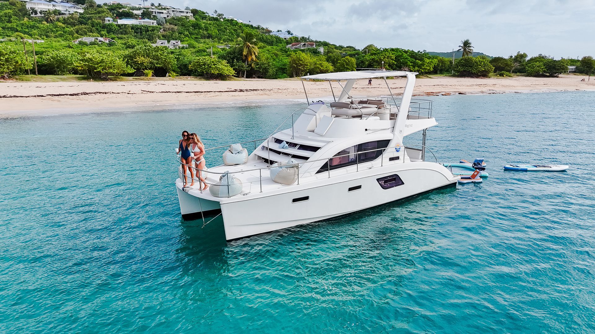 A white catamaran with people, turquoise water, and a beach in the background.