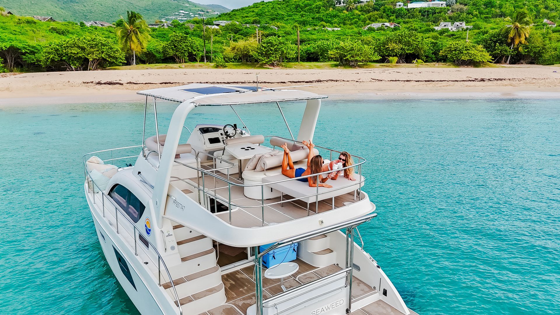 White yacht with people sunbathing on deck in turquoise water near a beach.