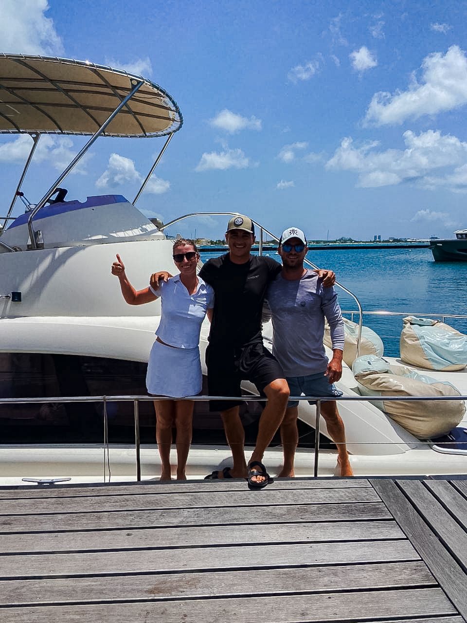 Three people smiling, posing on a wooden dock next to a white boat in sunny blue water.