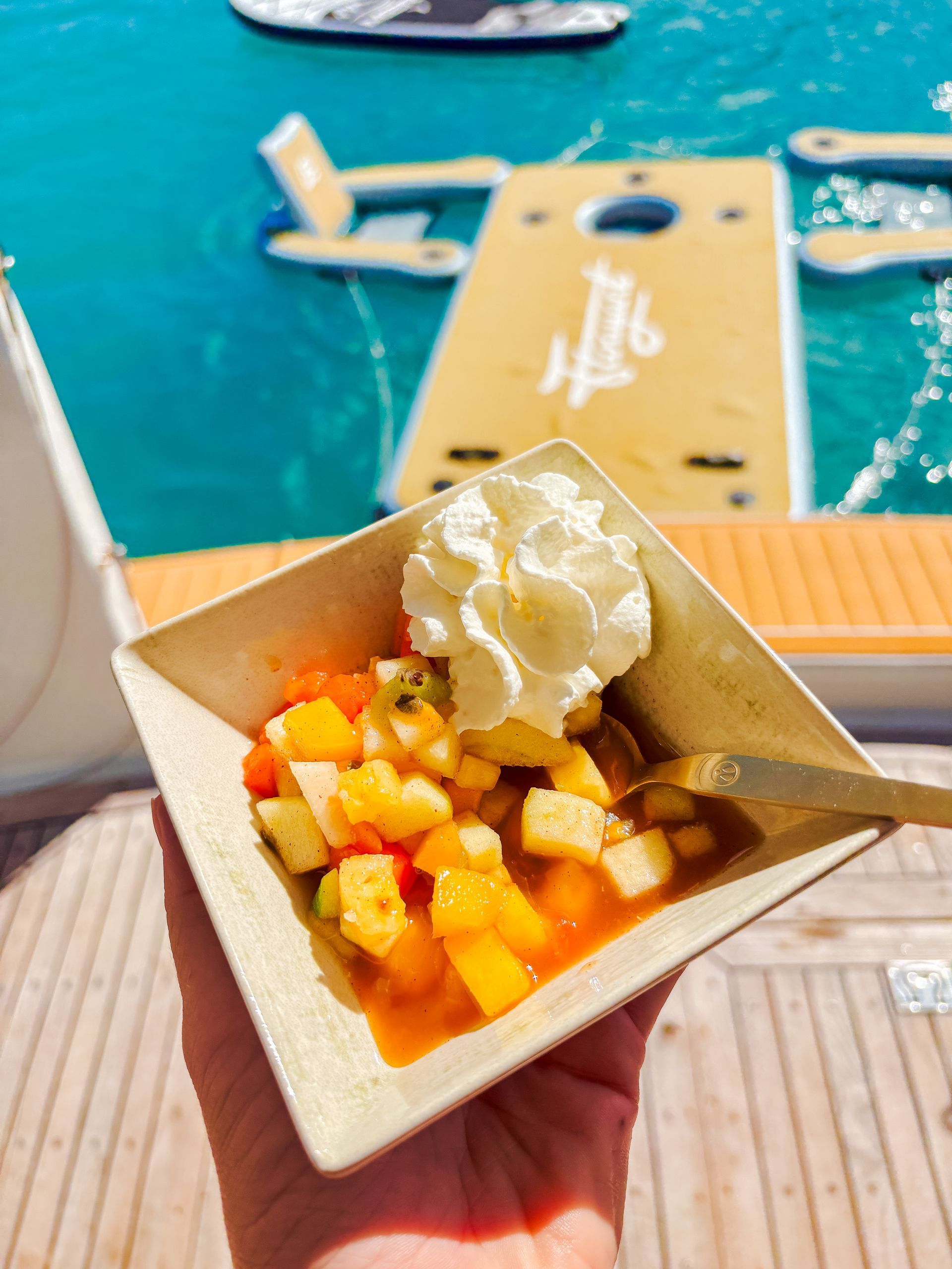 Hand holding a bowl of fruit salad with whipped cream, near turquoise water and a floating platform.
