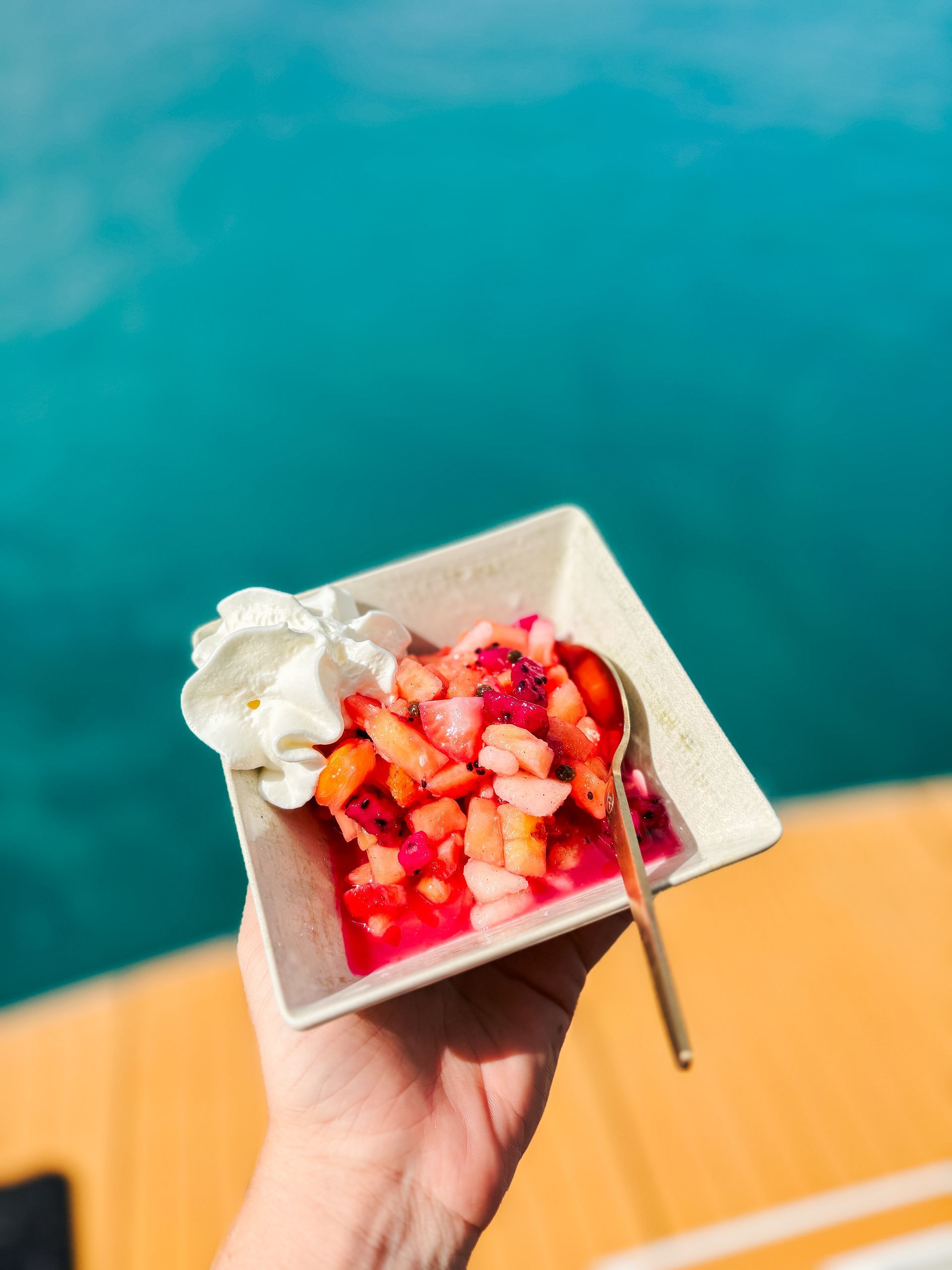 Hand holding a white square bowl with fruit salad, whipped cream, and gold spoon, against a blue water background.