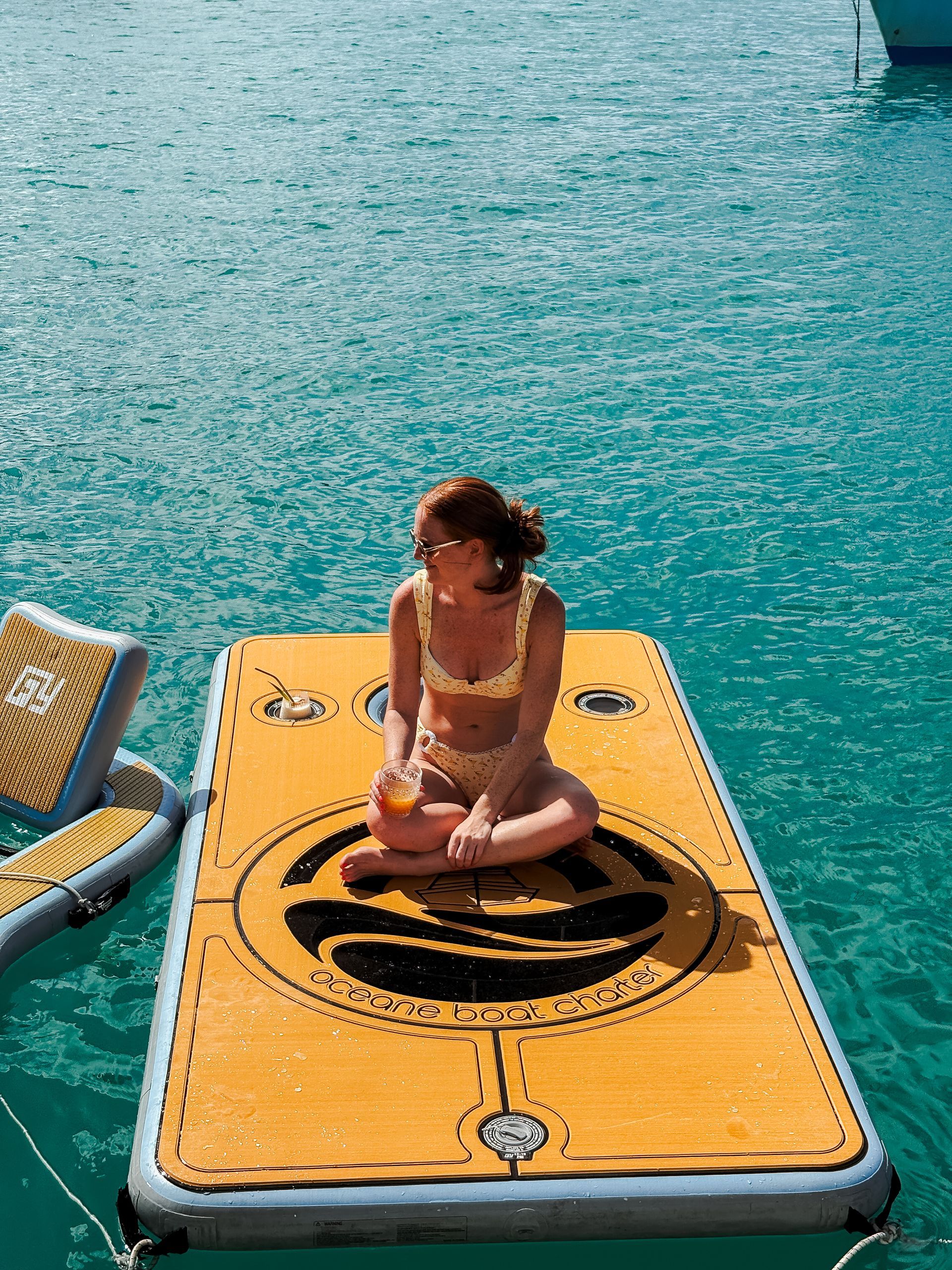 Woman in a bikini sits on a yellow floating mat in turquoise water, holding a drink.