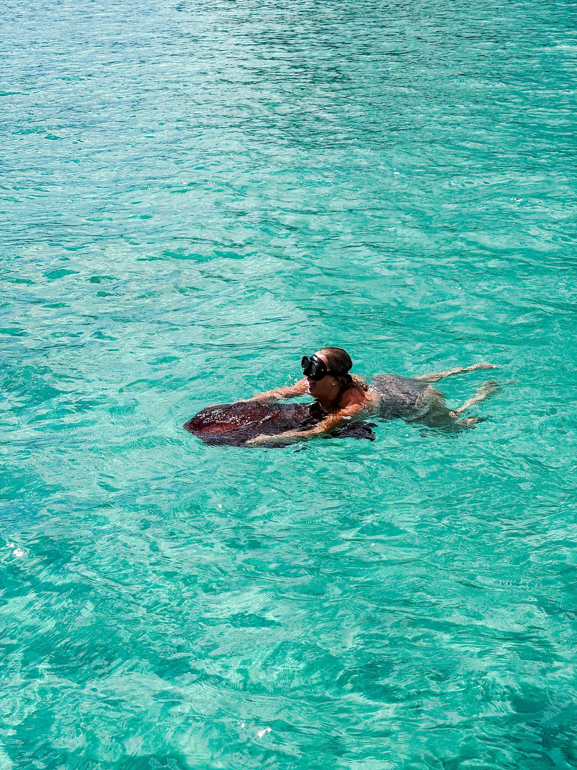 Person wearing snorkel gear, swimming with a maroon boogie board in turquoise water.