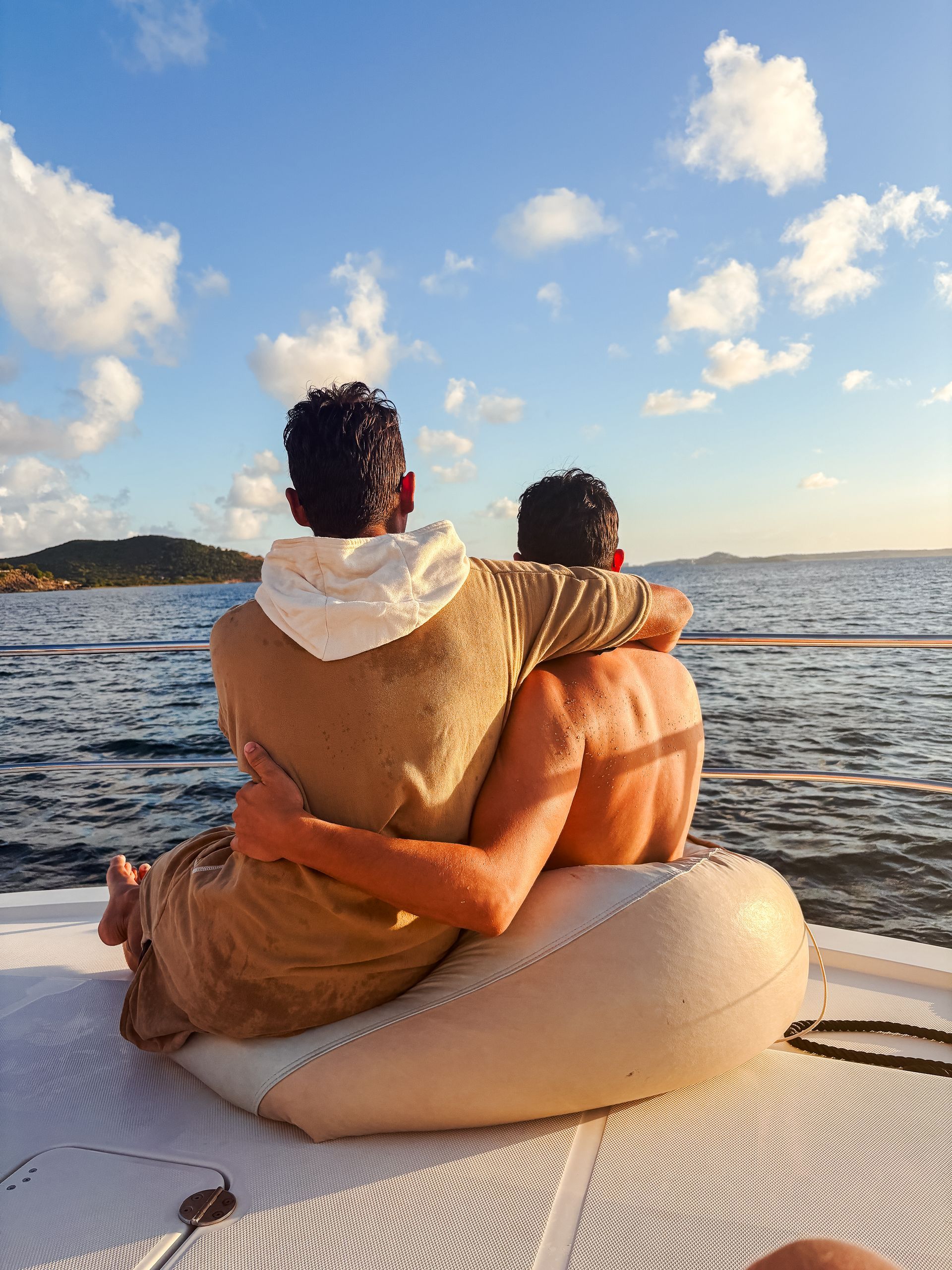 Two people embracing on a boat, looking out at the ocean under a blue sky with fluffy clouds.