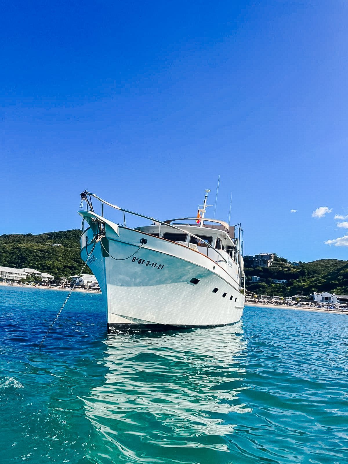 White yacht floating in turquoise water under a blue sky, with a green, hilly shoreline in the background.