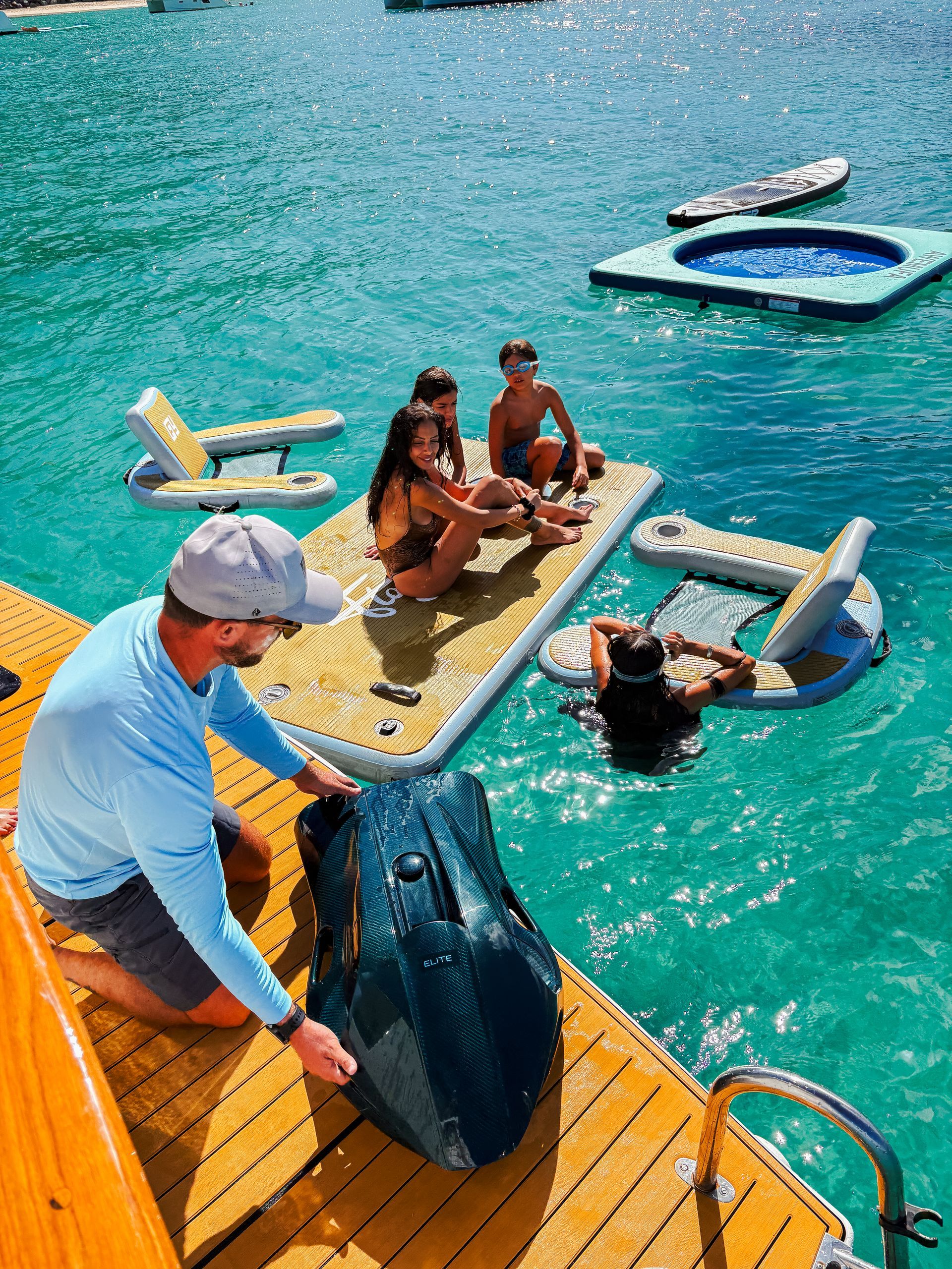 Man on dock preparing watercraft, people relaxing on inflatable platforms in turquoise water.